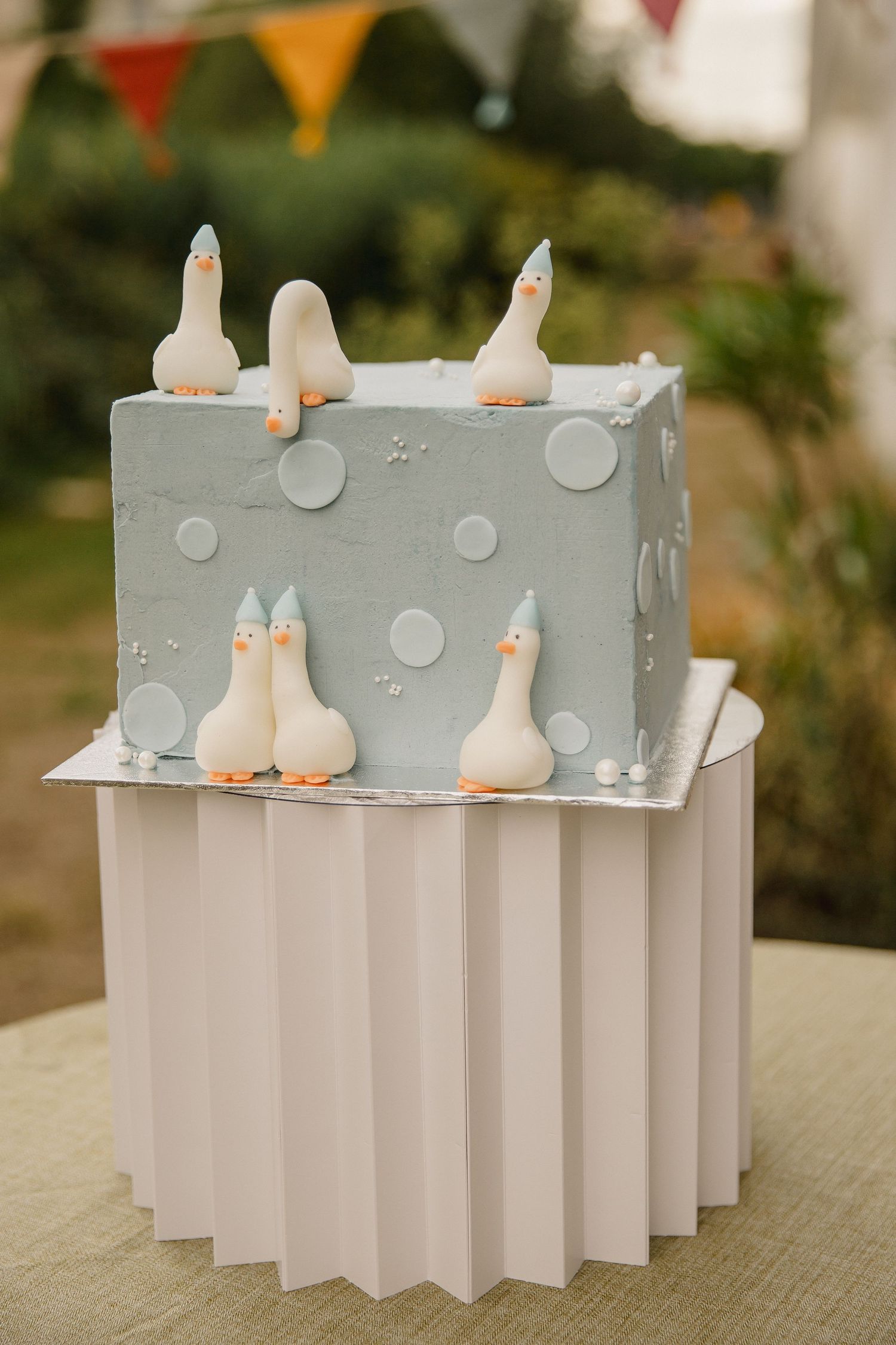 Square pale blue cake decorated with white polka dots and fondant geese, displayed on a pleated paper stand with bunting behind.