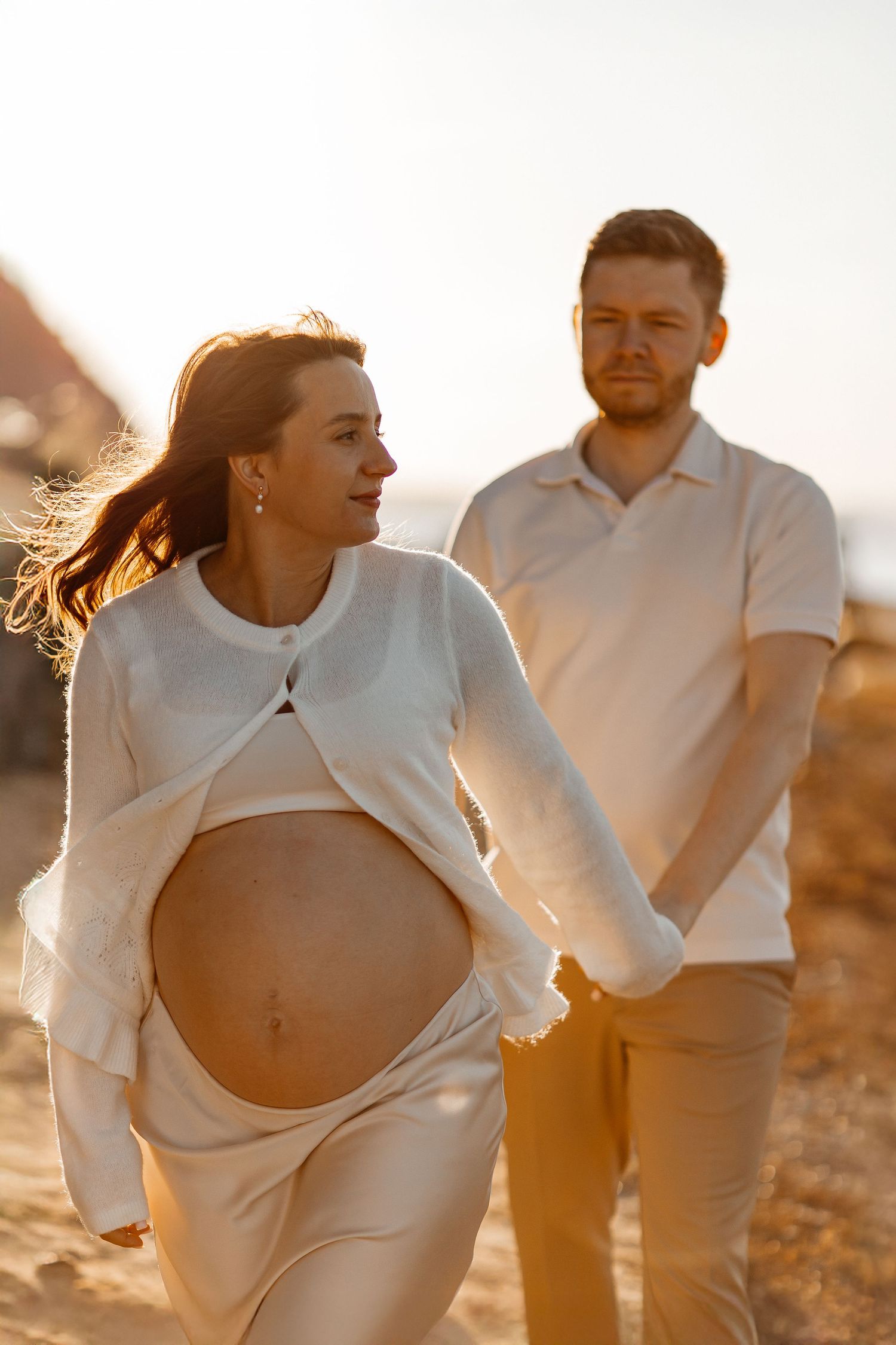 Pregnant woman in a white cardigan walks hand in hand with her partner on a sunlit beach at golden hour, her baby bump showing.