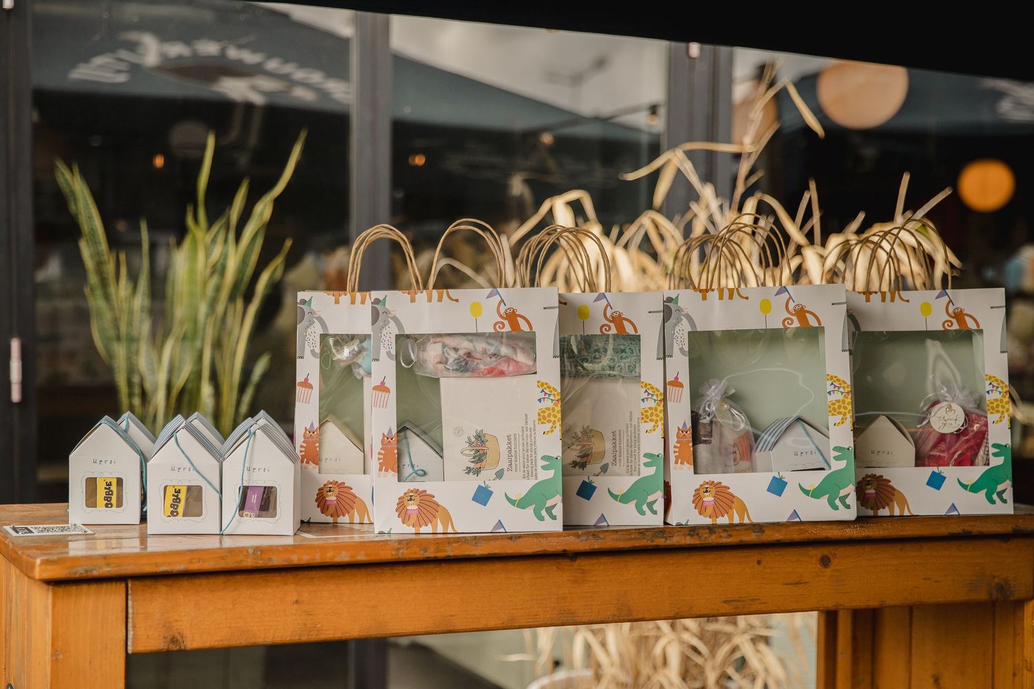 Gift boxes with dinosaur-print wrapping and clear windows displaying small house-shaped ornaments, arranged on a wooden shop table.