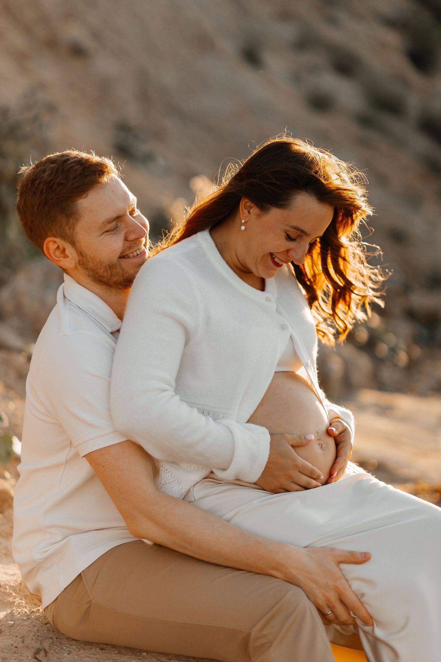 Smiling expectant couple embracing outdoors at golden hour, the woman cradling her baby bump while the man hugs her from behind.