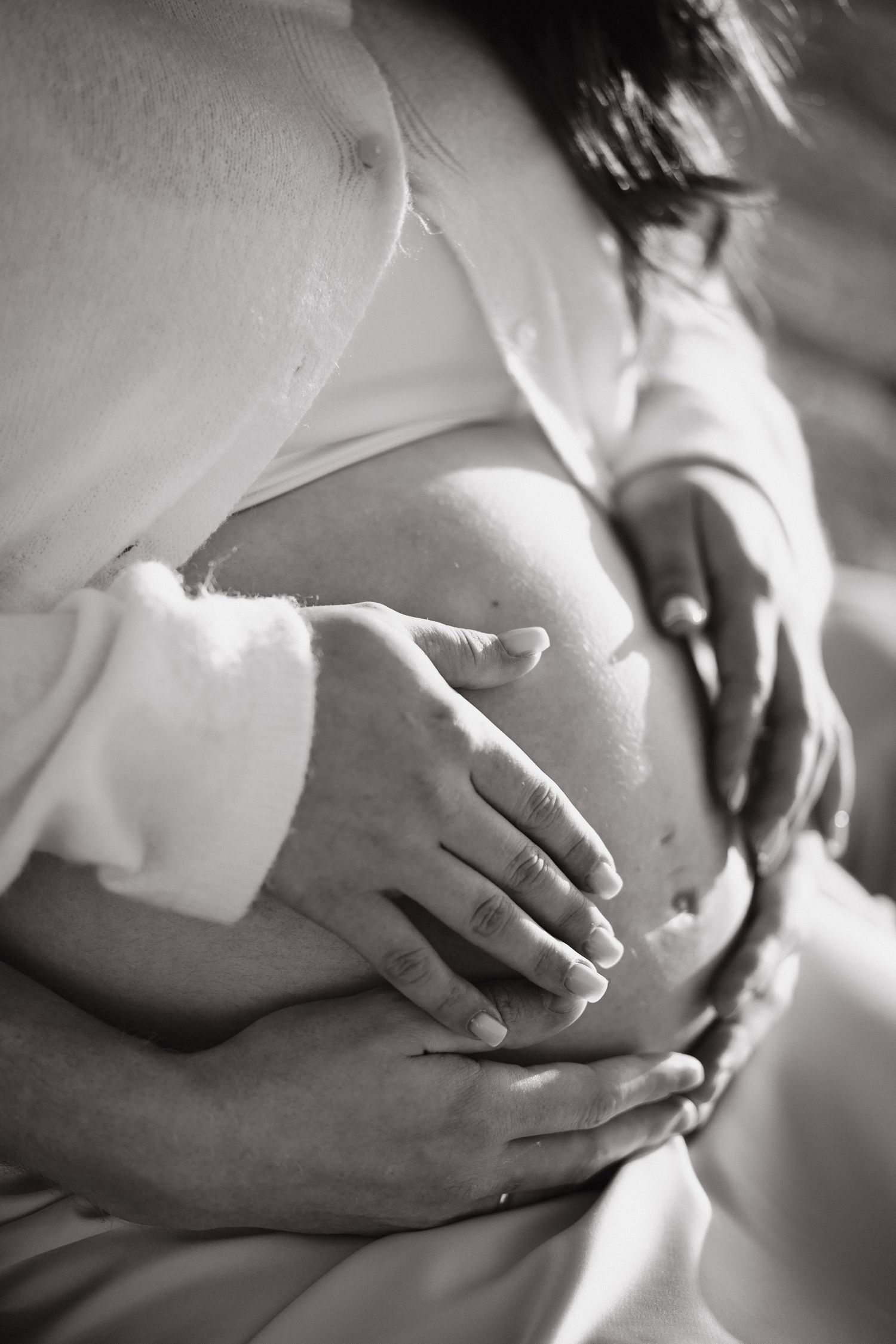 Black and white photo of several hands gently resting on a pregnant woman's bare belly, capturing a tender maternity moment.