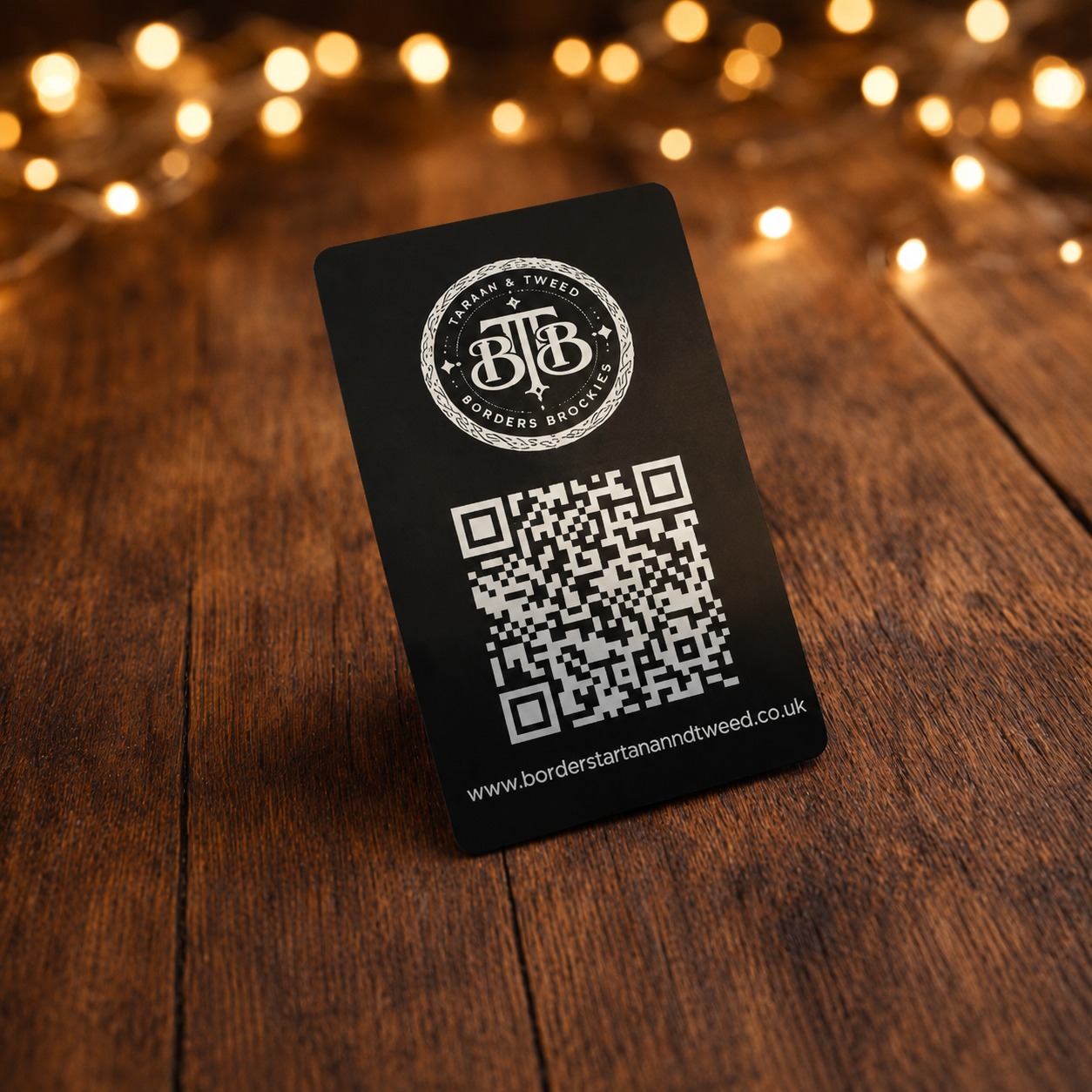 Black Borders Tartan and Tweed business card with circular logo and QR code, standing on a rustic wooden table with warm fairy lights.