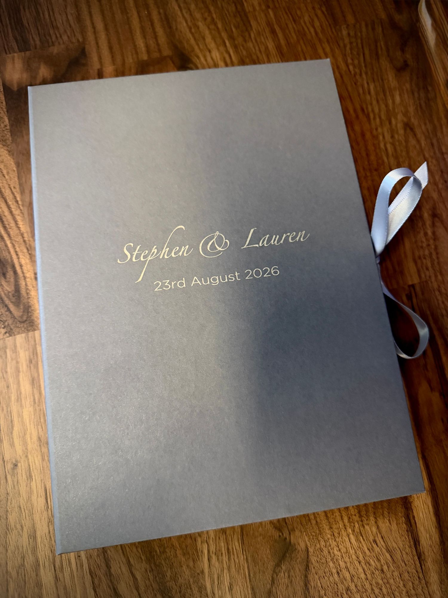 Wedding invitation booklet for Stephen and Lauren dated 23rd August 2020 with blue ribbon on wooden surface.
