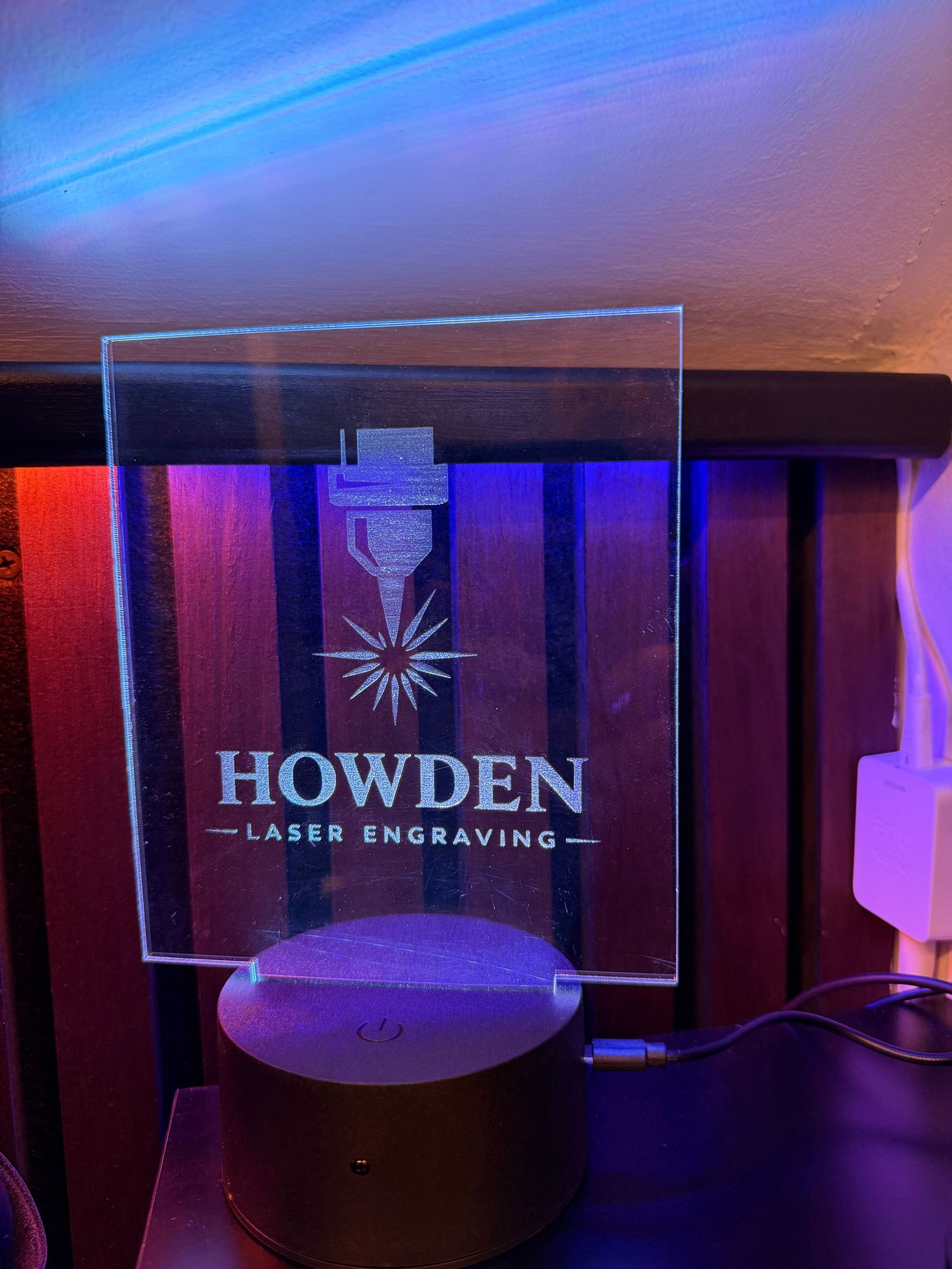 Laser engraving machine illuminated with blue and purple lighting creates glowing patterns in a dark workshop setting.