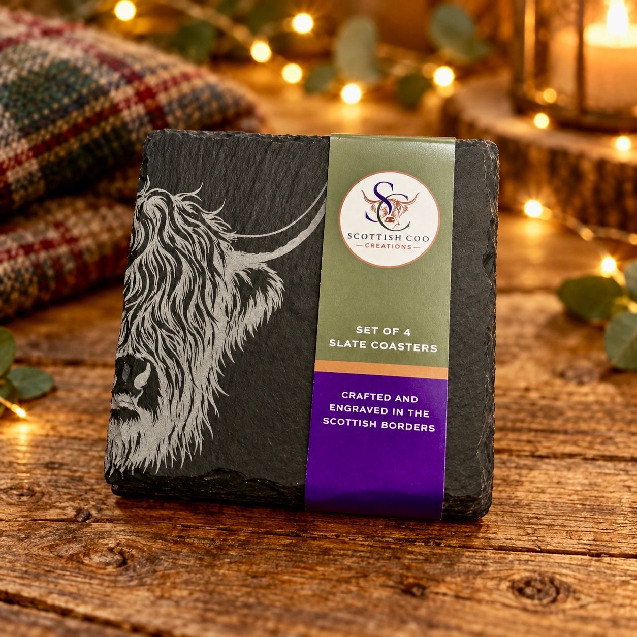 Set of 4 Scottish Coo Creations slate coasters engraved with a Highland cow, in packaging on a wooden table with festive lights.