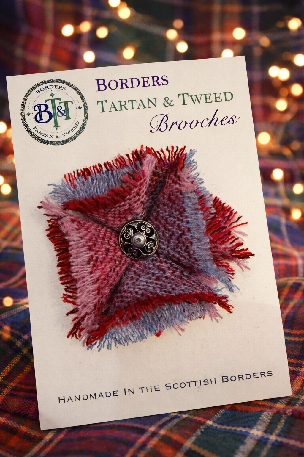A handmade Scottish tartan and tweed brooch with red and blue frayed edges displayed on a product card with bokeh lights.