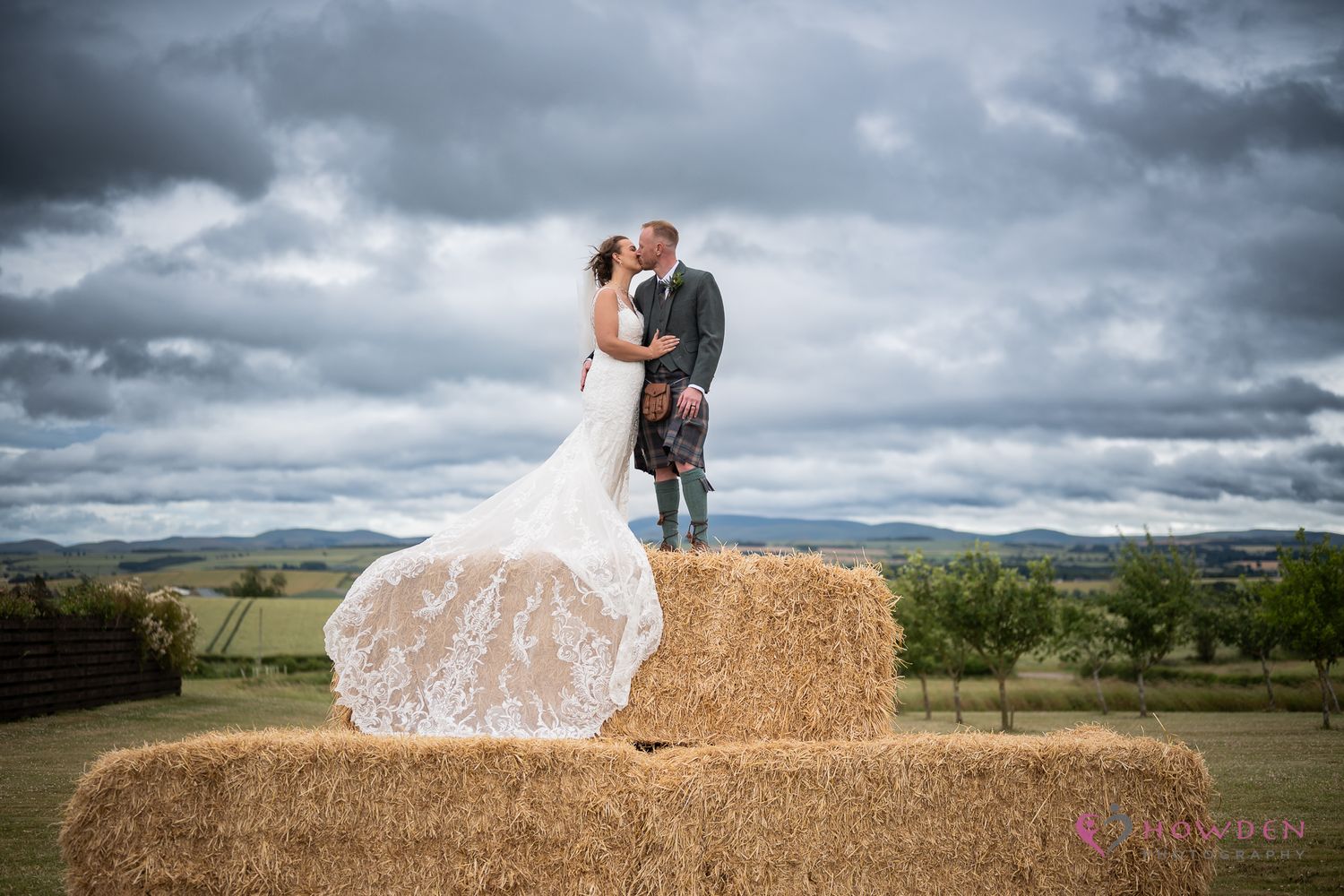 Bride in a lace wedding gown and groom in a kilt kiss atop stacked hay bales under a dramatic cloudy sky in the countryside.