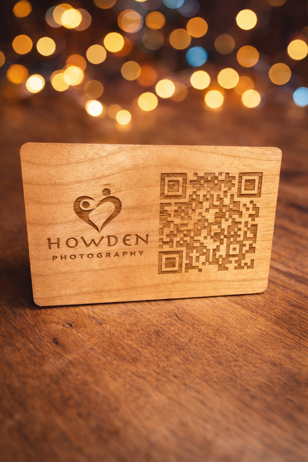 Wooden business card with Howden Photography logo and QR code on wooden surface with bokeh lights in background.