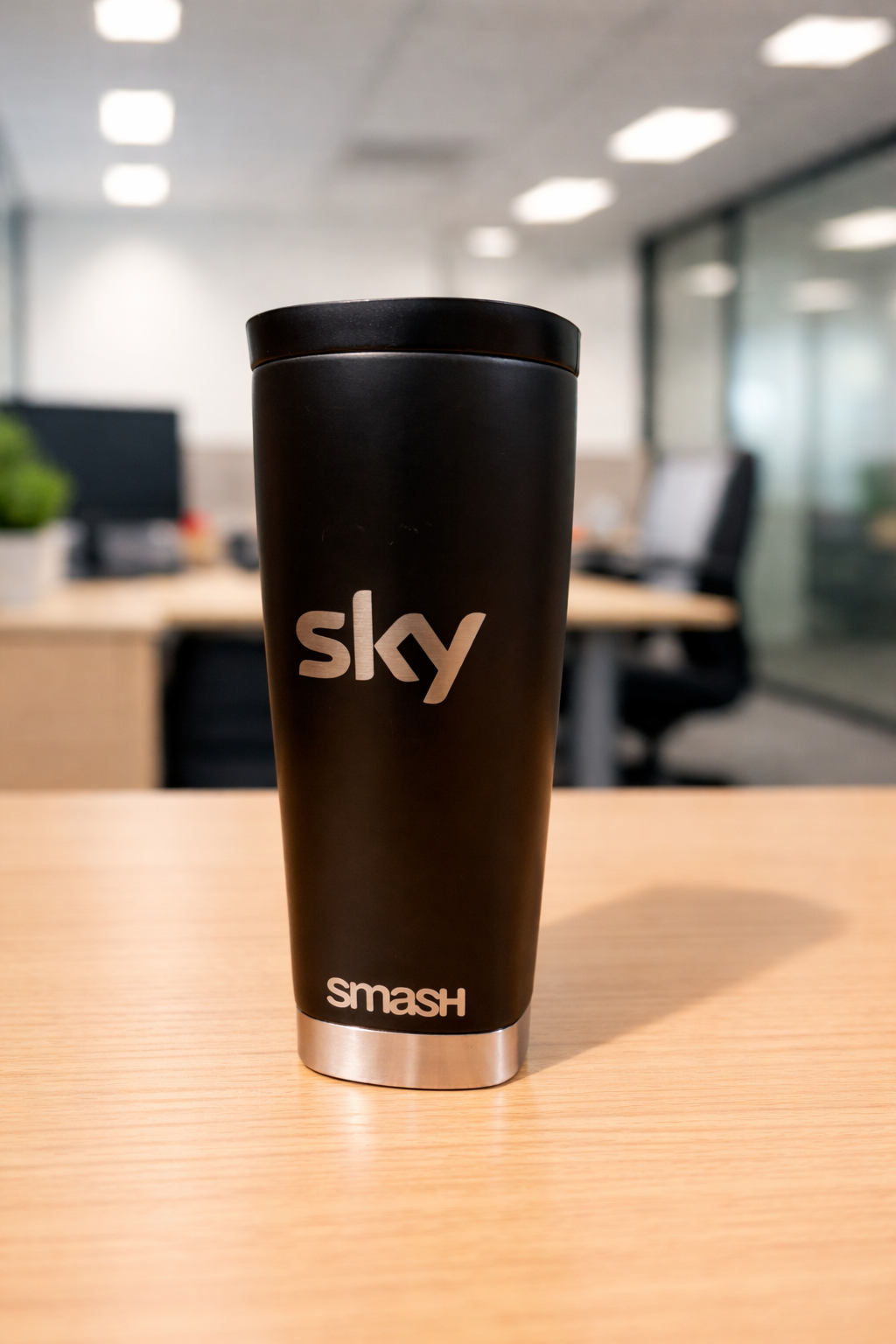 A black Sky Smash branded coffee cup shown from multiple angles on a wooden desk in an office setting.