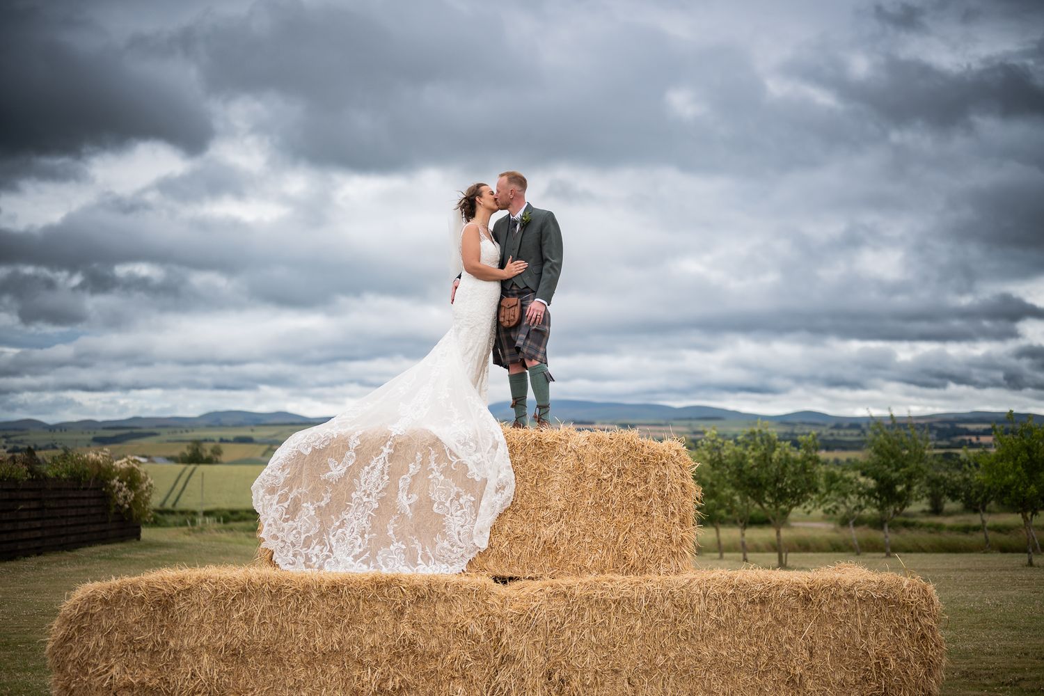 Bride in a lace wedding gown and groom in a kilt kiss while standing on stacked hay bales with rolling countryside and a cloudy sky behind.