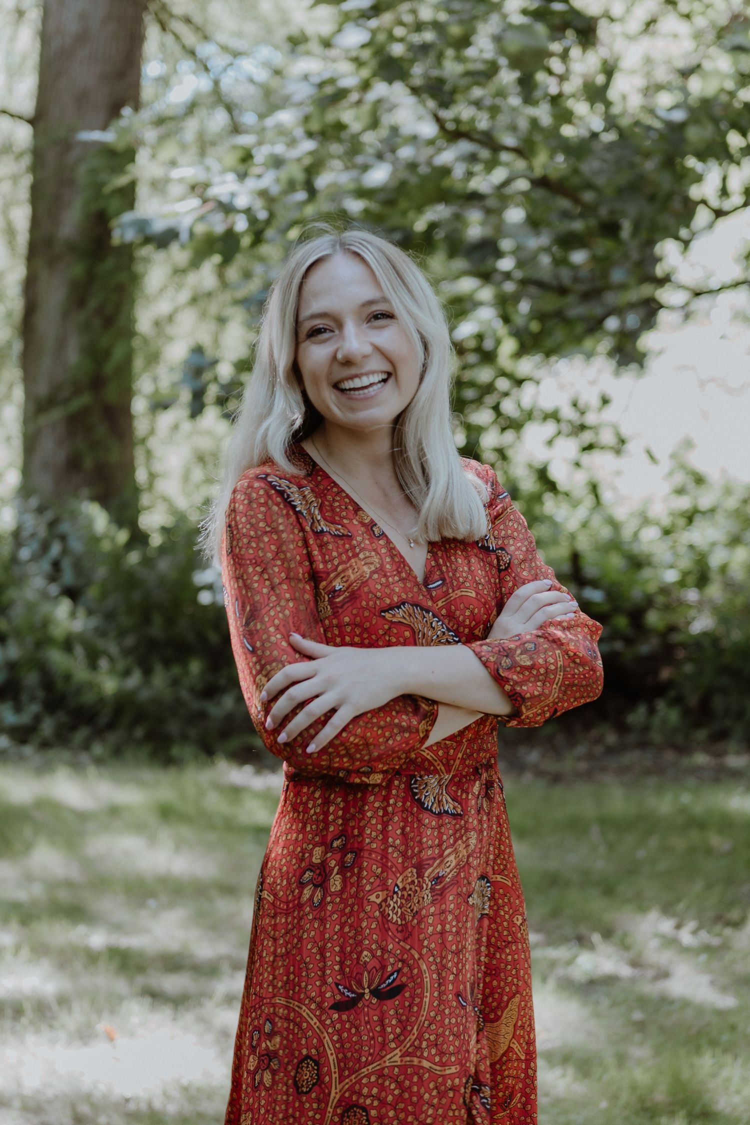 A cheerful portrait of someone in a red patterned dress standing in a sunlit garden.