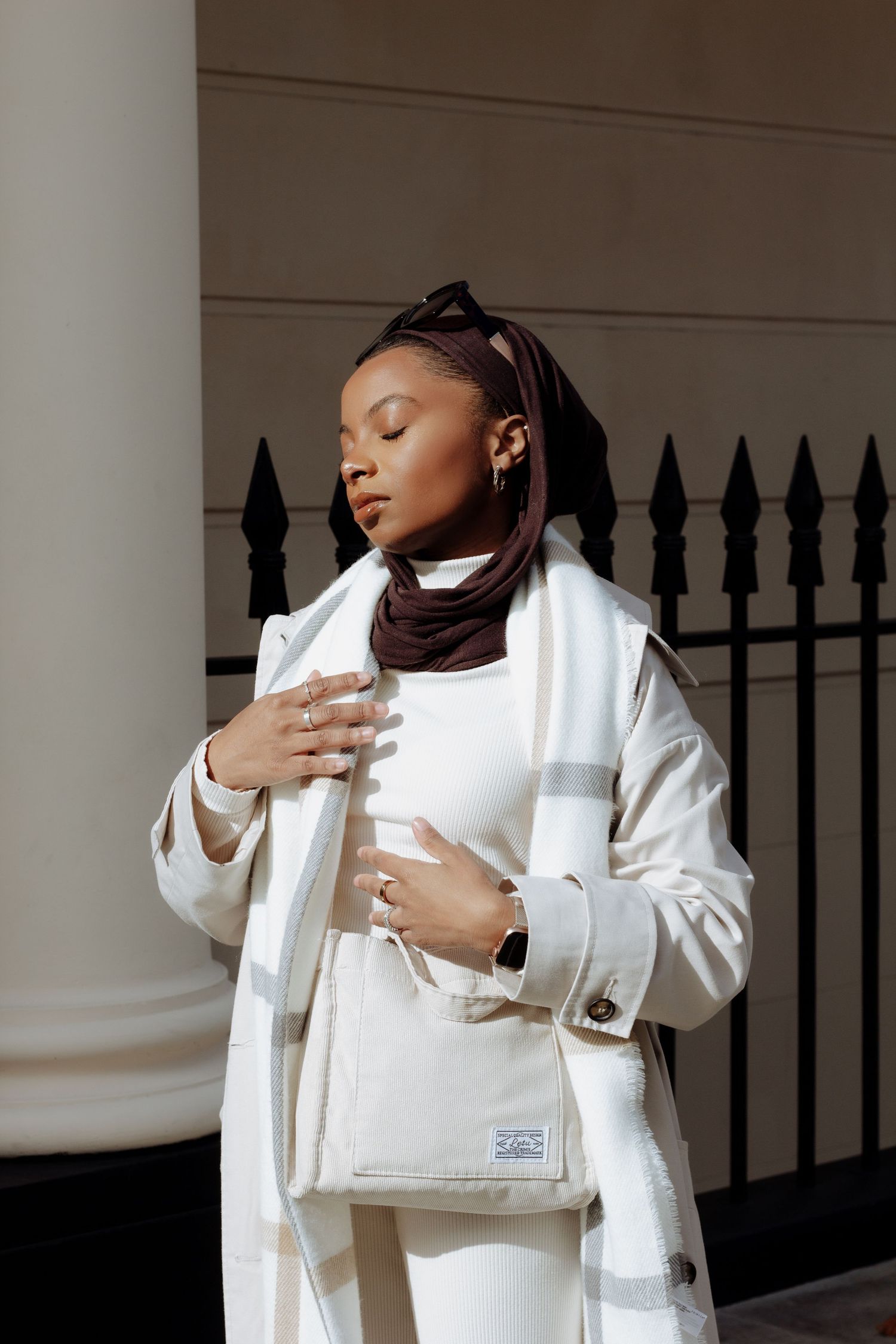 Stylish winter fashion with brown hijab, white coat and designer handbag against iron fence backdrop.