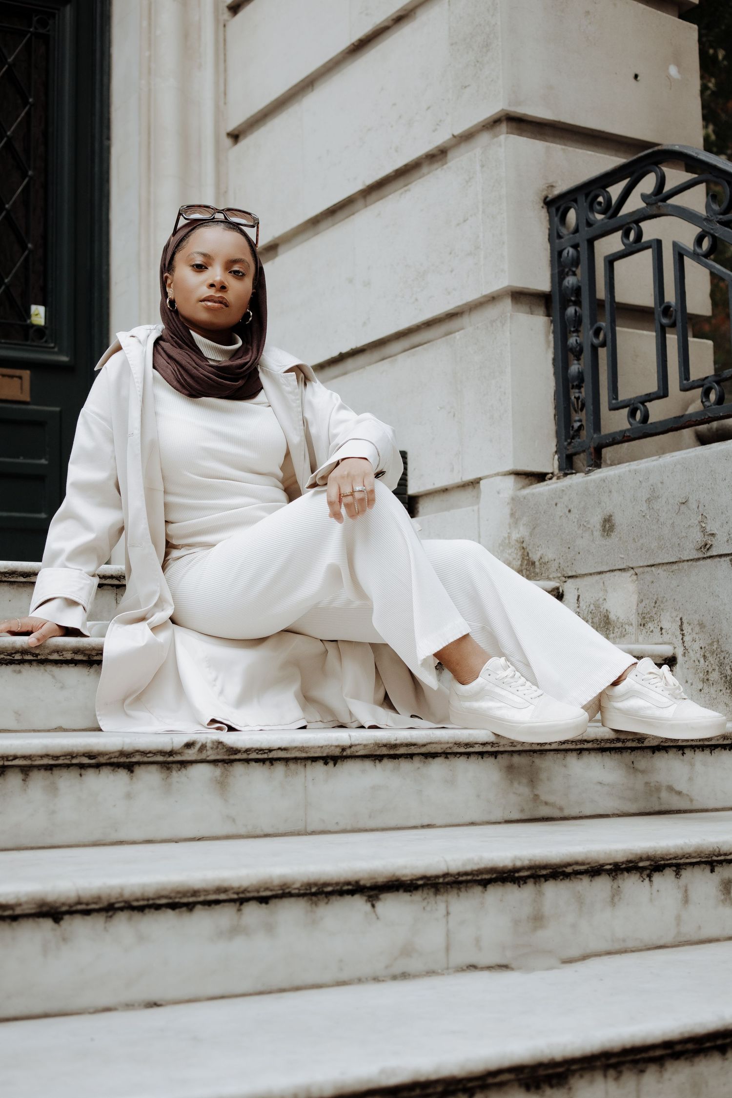 Person in all-white outfit with brown headscarf sitting on concrete steps of a building entrance.