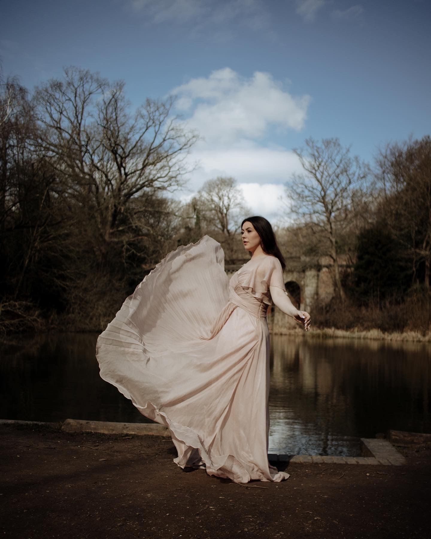 Flowing pink dress billows dramatically by a serene pond surrounded by bare winter trees on a cloudy afternoon.