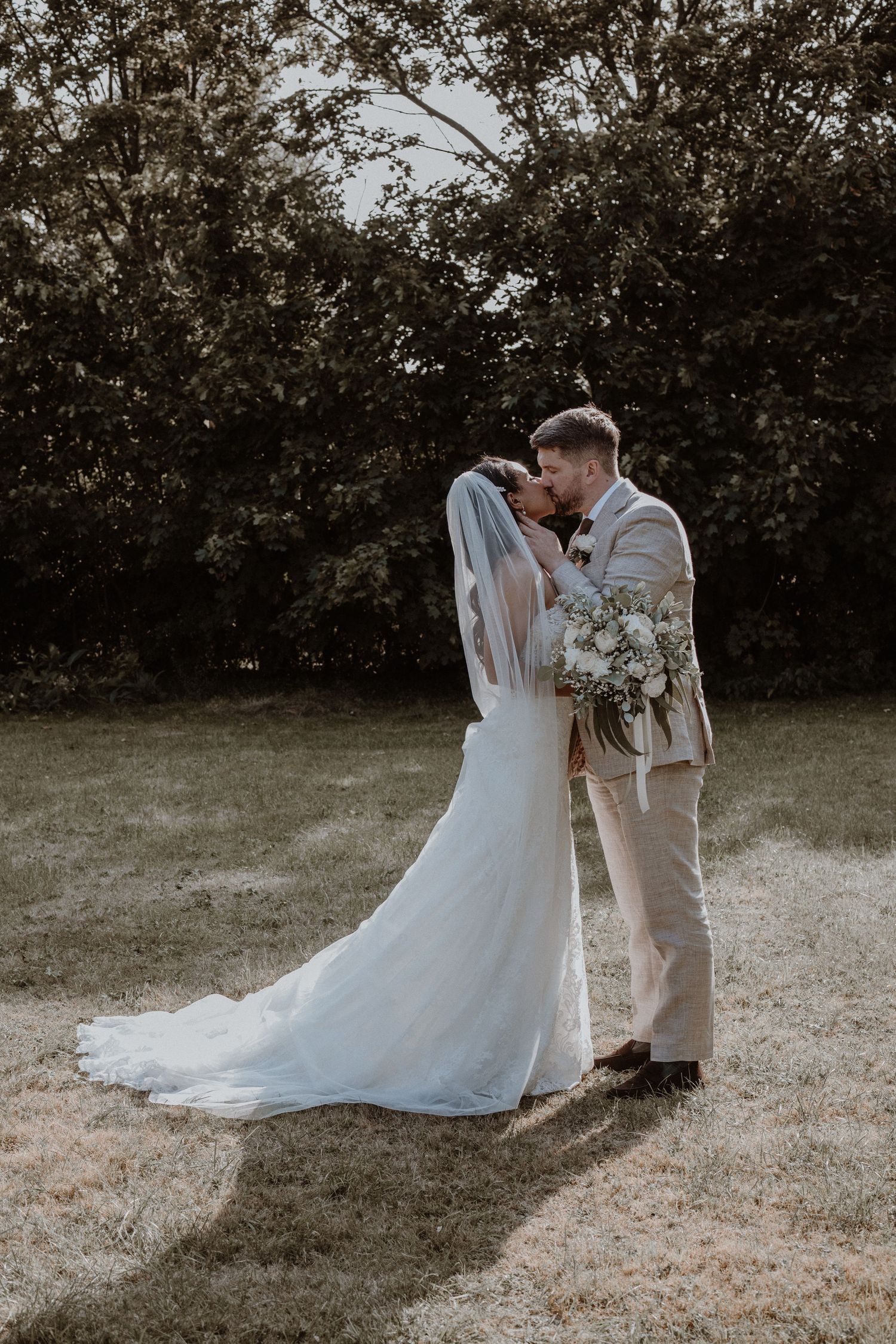 Romantic wedding couple sharing intimate moment outdoors with flowing white dress and delicate bouquet at sunset.