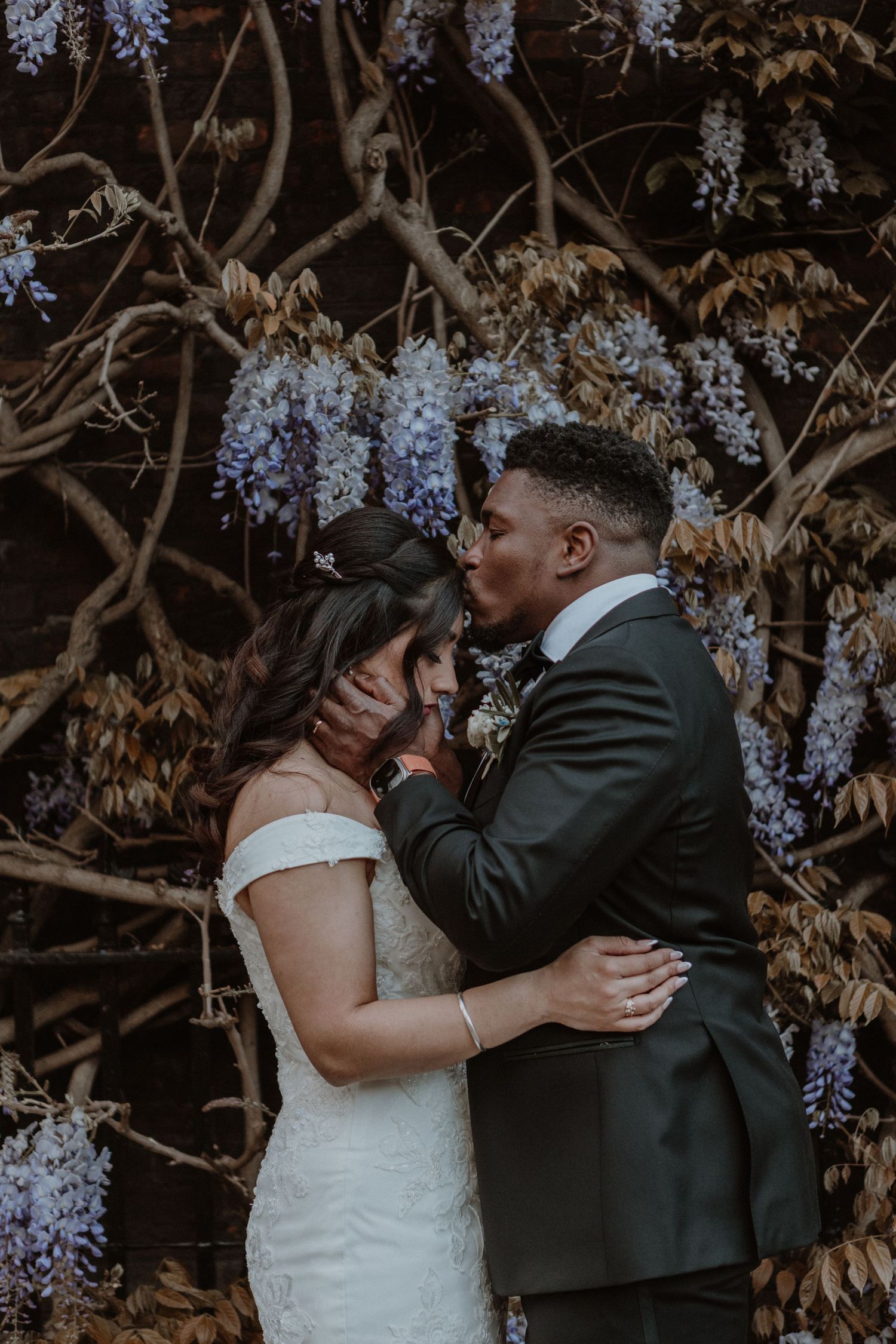 Newlywed couple shares intimate moment under blooming purple wisteria flowers at outdoor wedding venue.