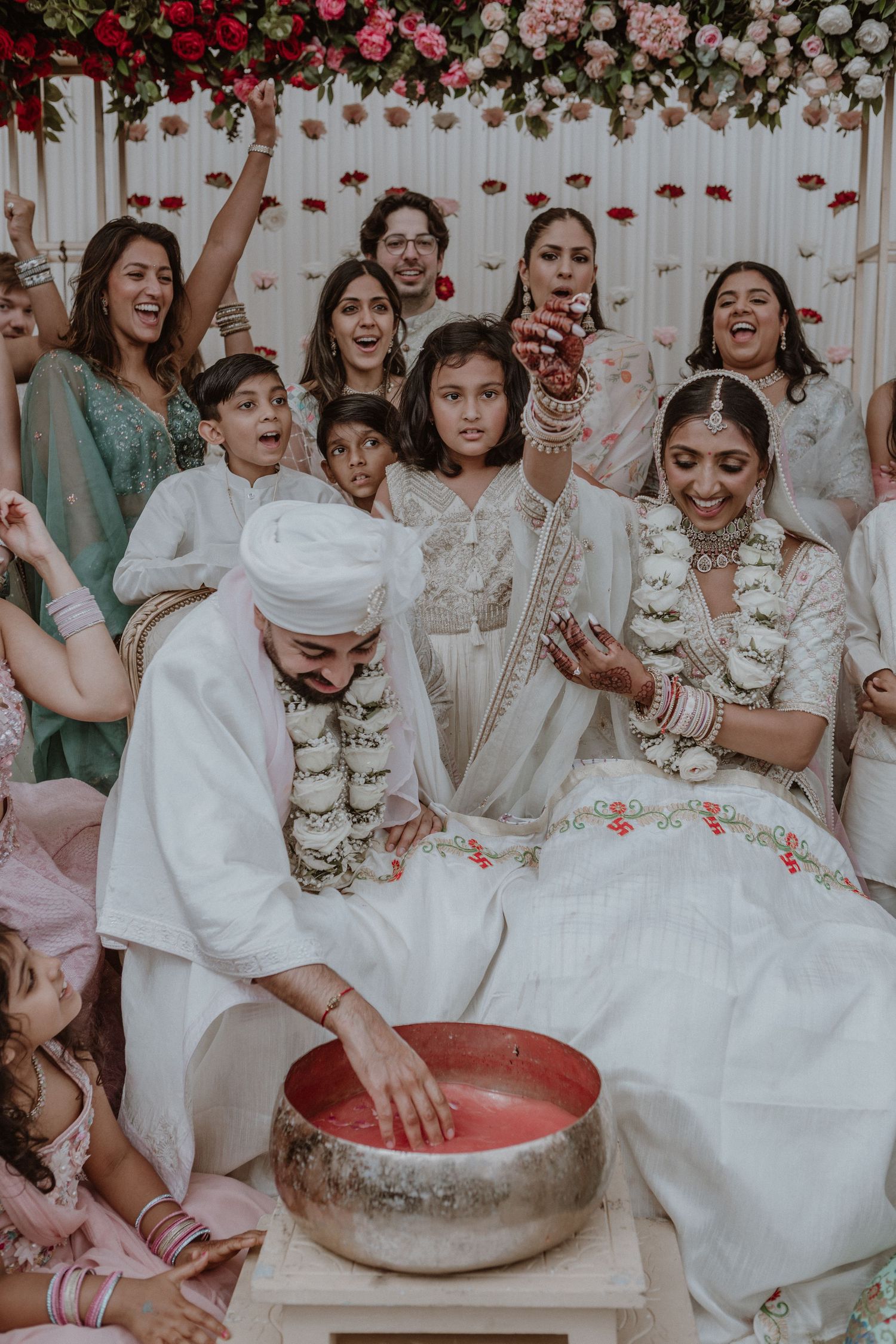 Festive traditional Indian wedding celebration with floral decorations and silver bowl ritual in progress.