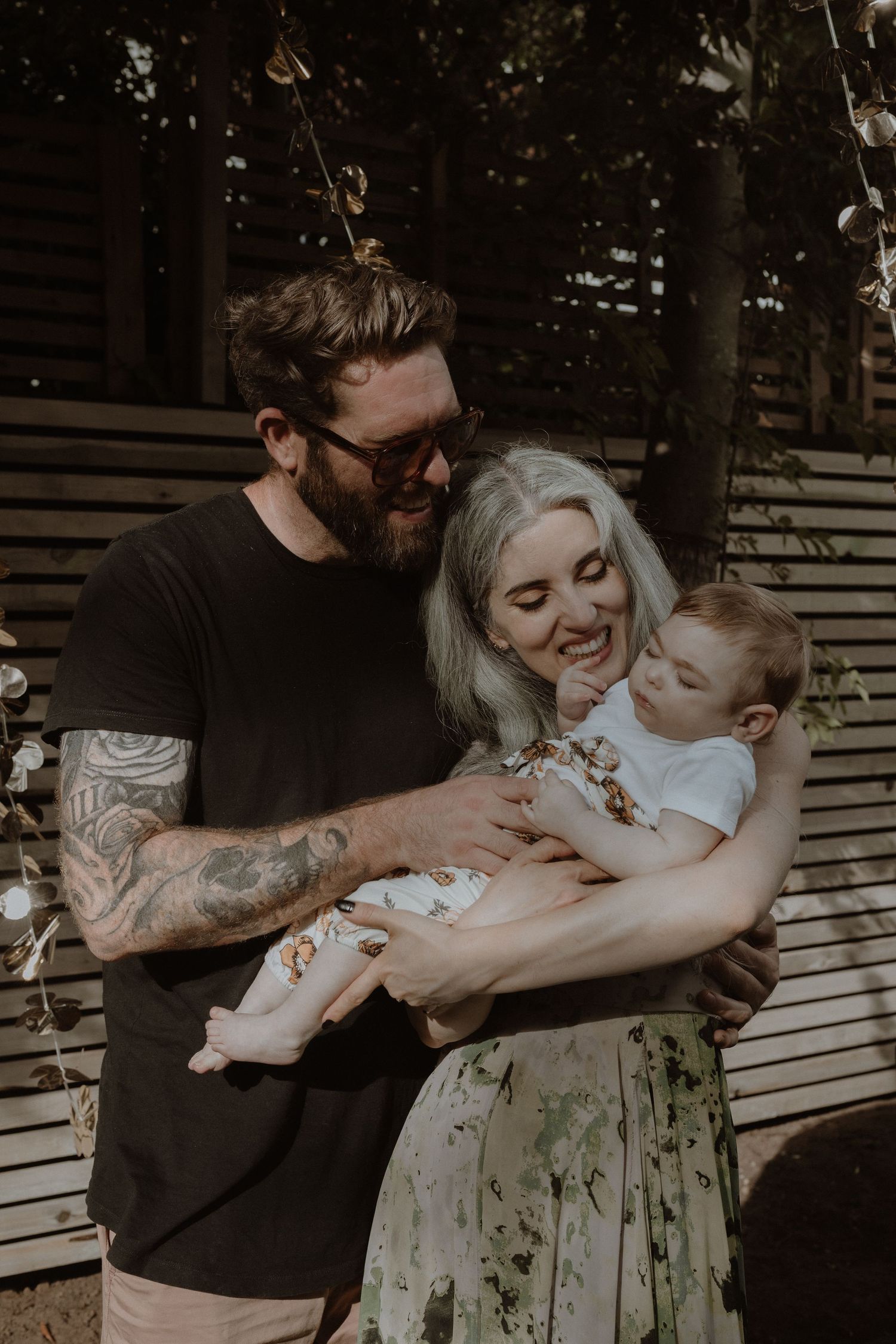 A couple shares a tender moment with their baby outdoors, smiling and embracing in warm natural lighting.