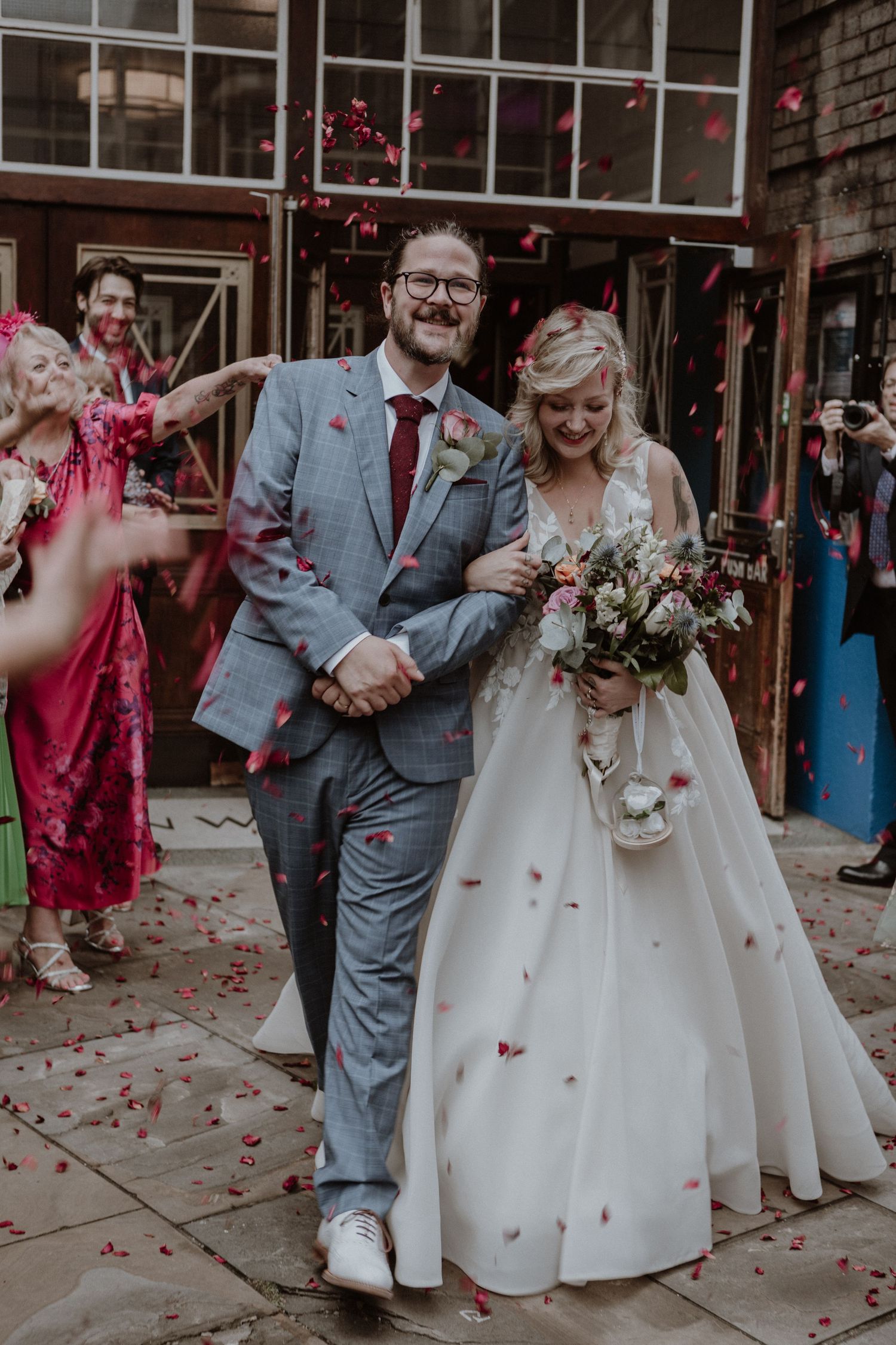A joyful wedding couple walking under rose petal confetti outside a brick building.