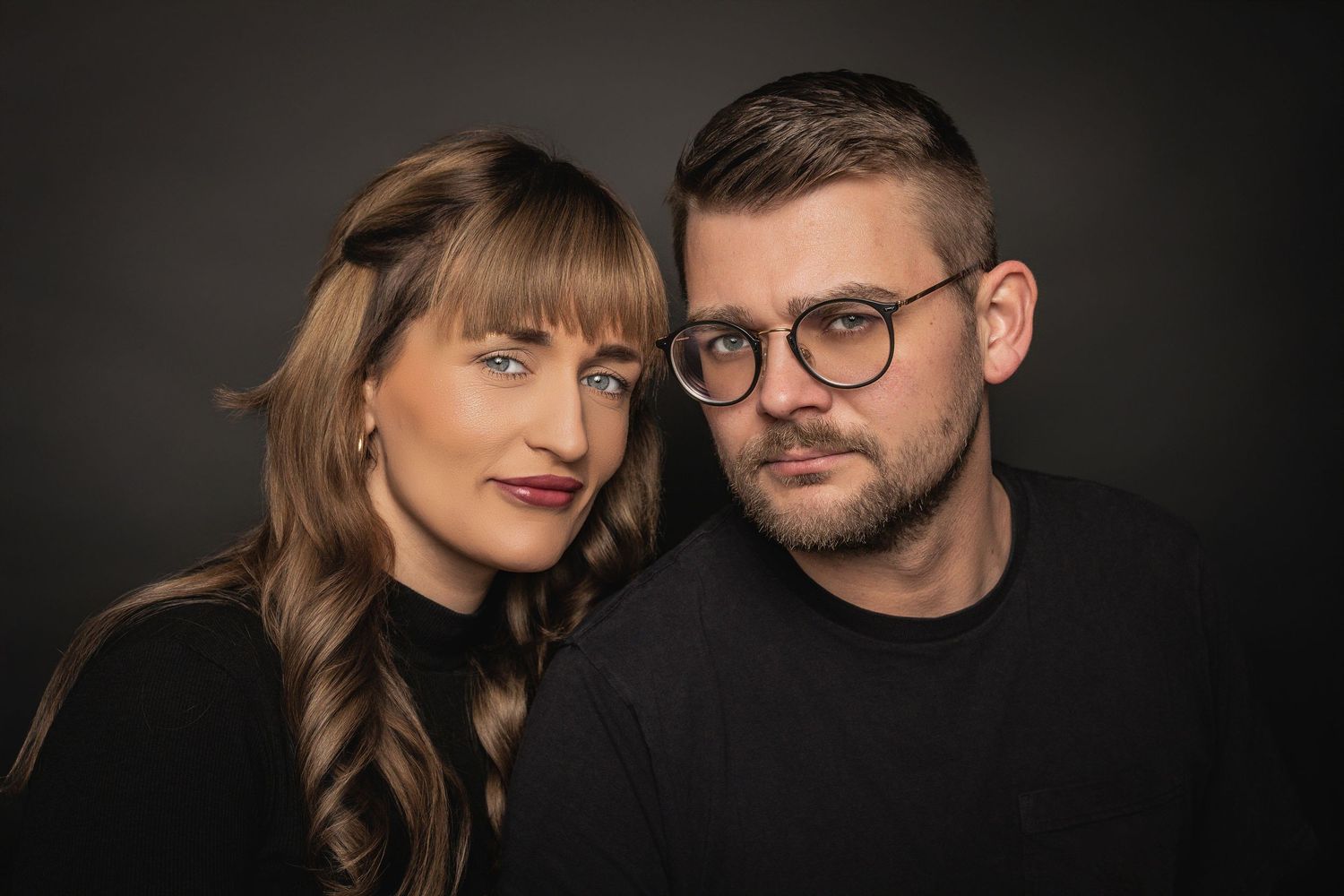 Artistic black and white studio portrait of two people against dark background with dramatic lighting.