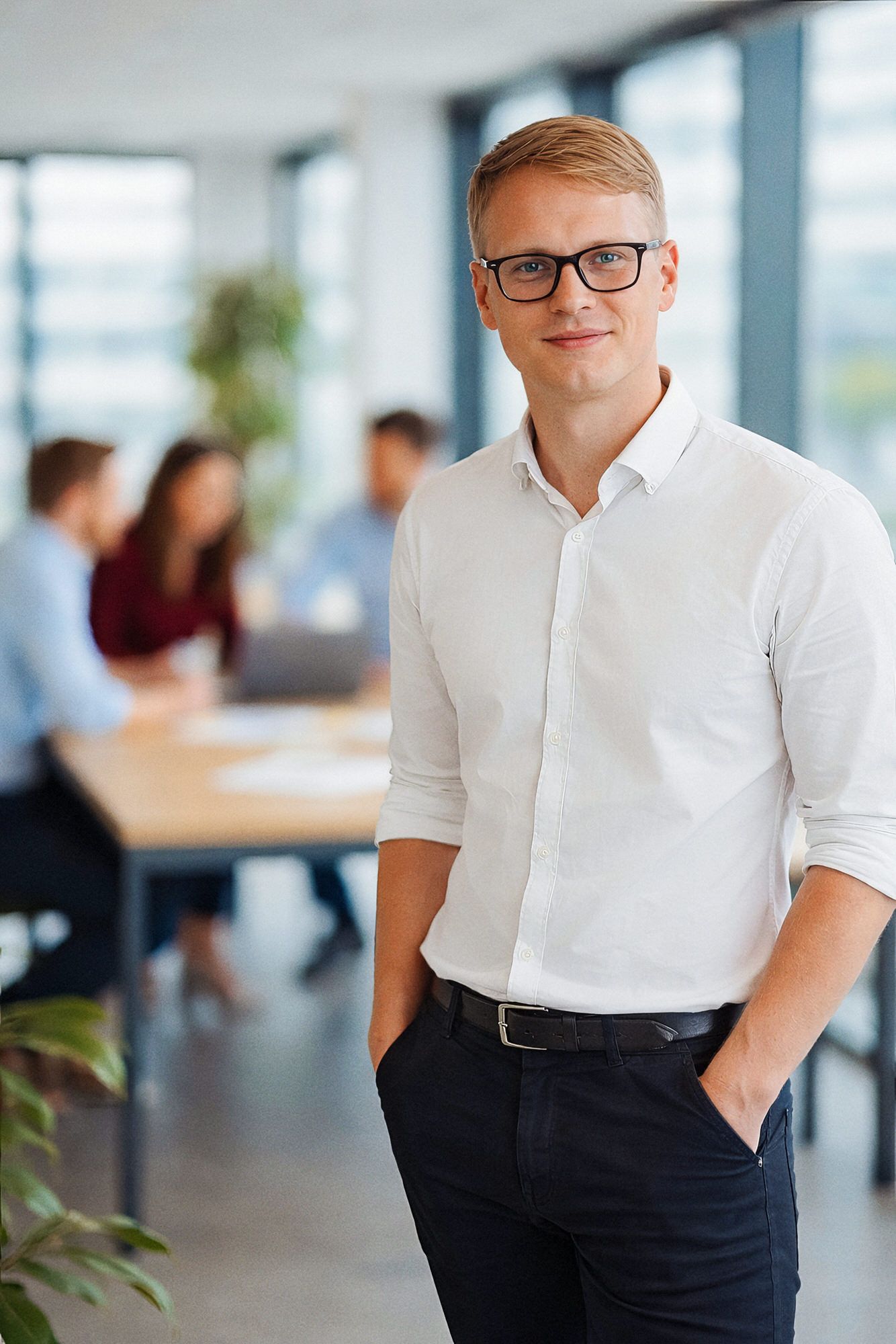 Professional in a white shirt and dark pants stands in a modern office with coworkers in the background.
