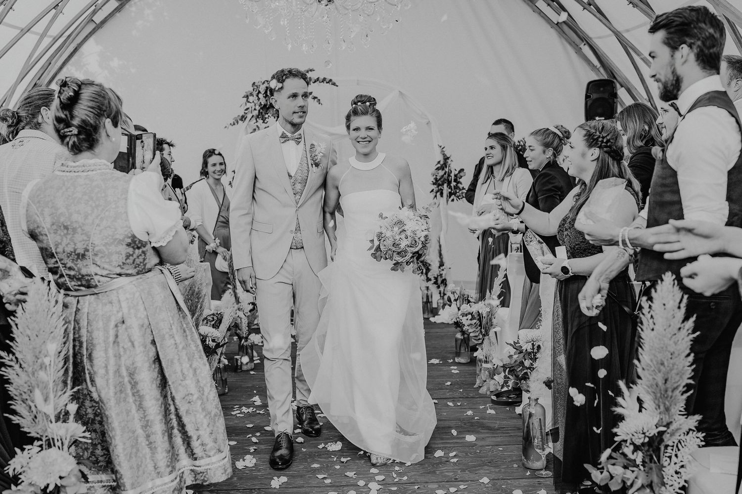 Black and white wedding photo showing guests throwing confetti over a couple during an outdoor celebration.