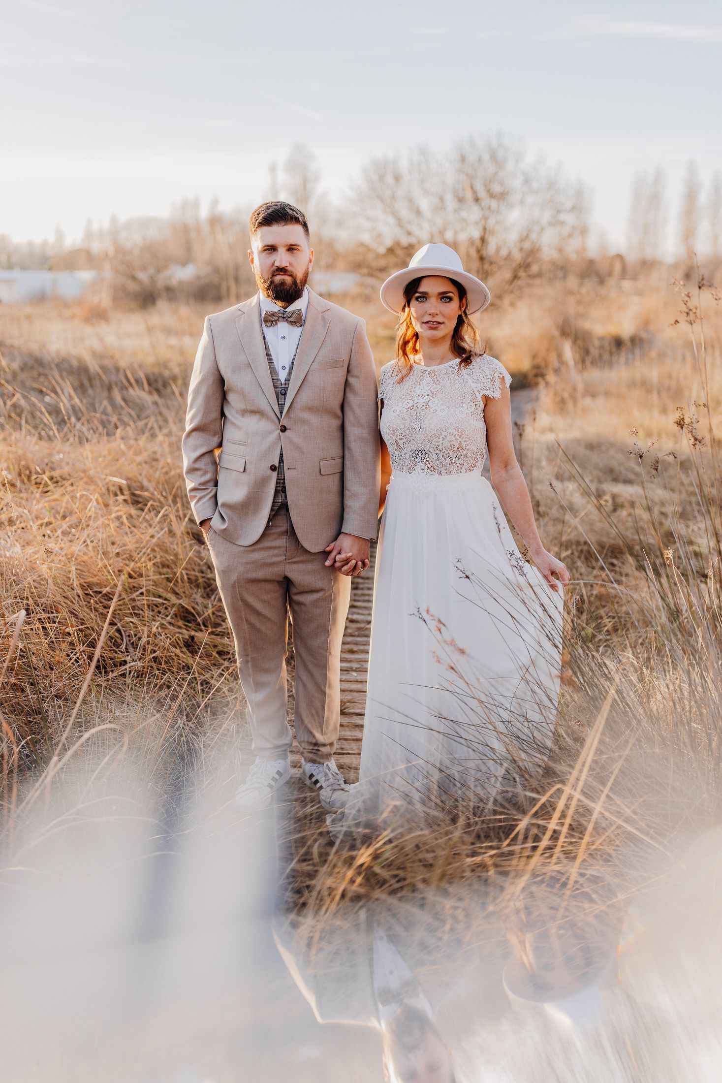 A couple in wedding attire poses together in a golden field of tall grass during sunset.