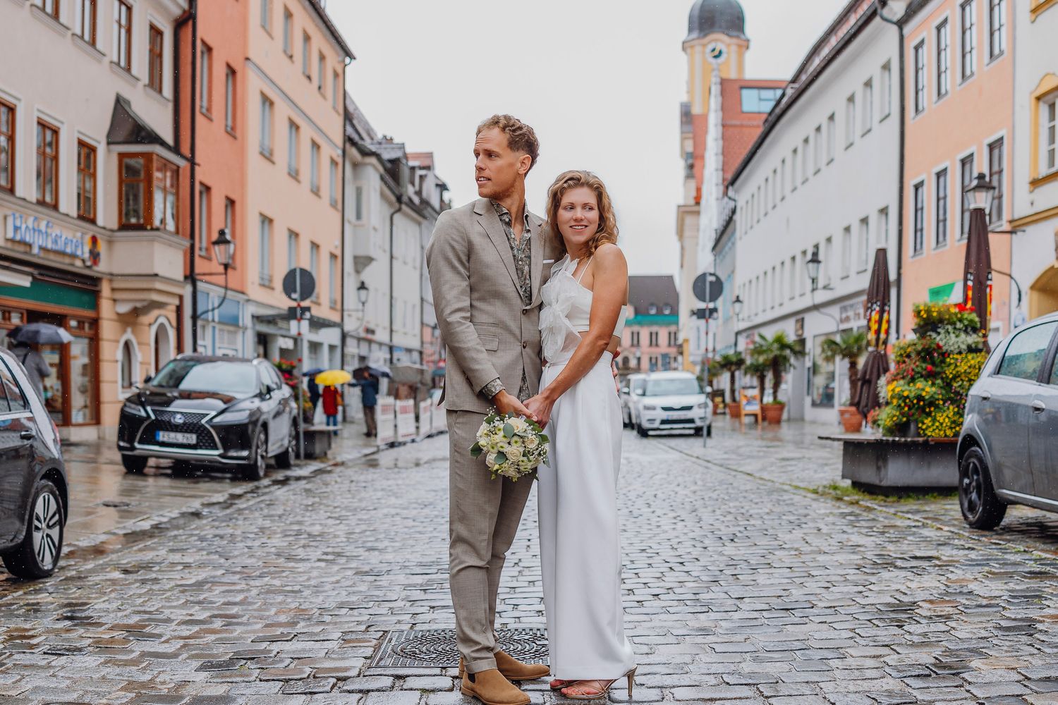 A couple in wedding attire stands on a cobblestone street in a charming European town with a church spire in the background.