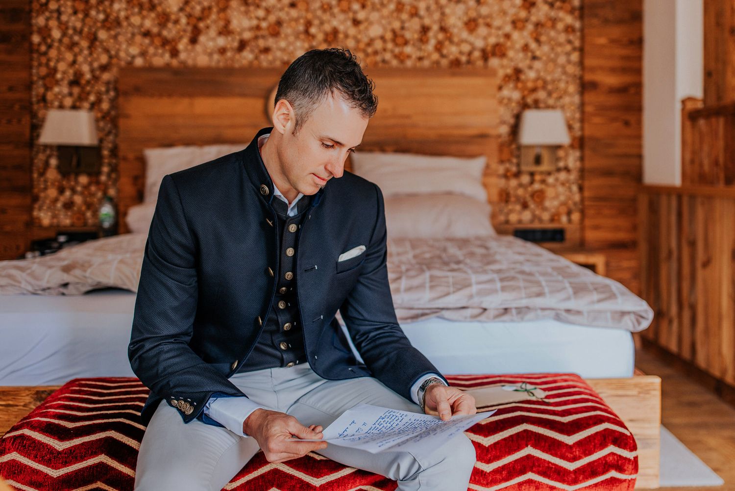 Person in traditional Austrian jacket sits on red and white patterned blanket in cozy wooden cabin bedroom.