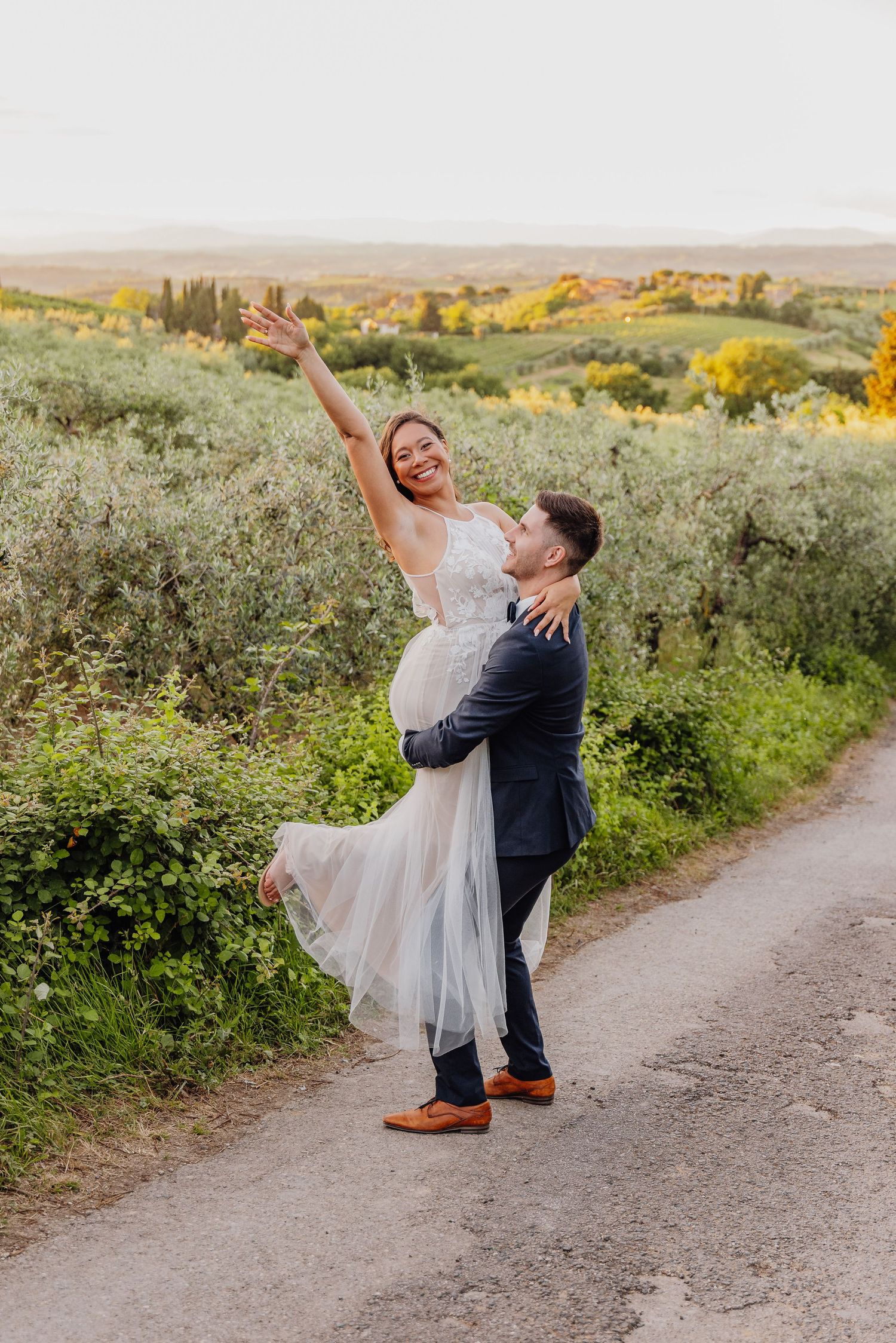 A couple in formal attire dances playfully on a dirt path surrounded by lush greenery and golden wildflowers.