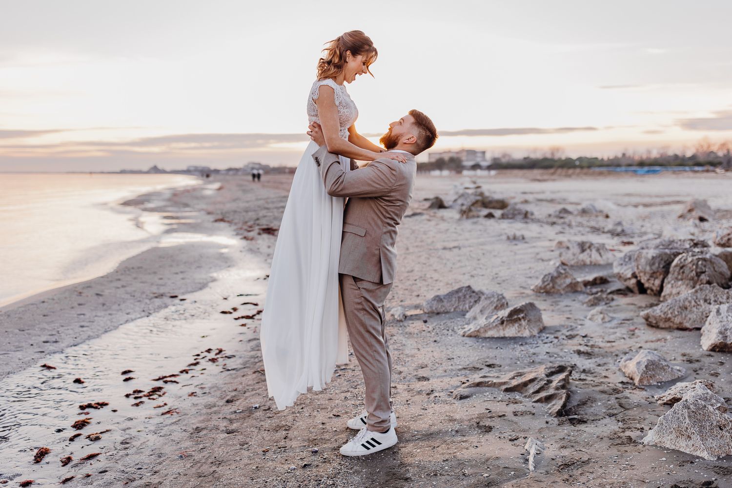 A romantic couple embraces on a rocky beach at sunset, with the bride wearing a flowing white dress and groom in grey.
