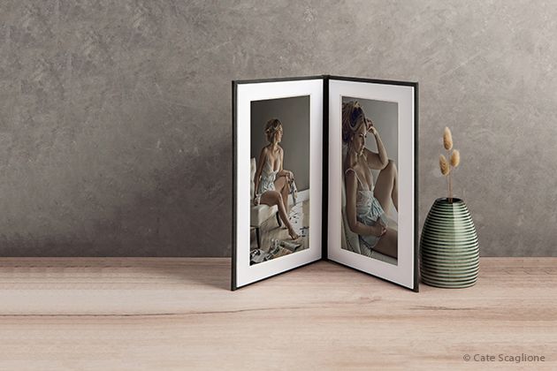 A framed double photo display stands beside a green ceramic vase on a beige surface against a textured gray wall.