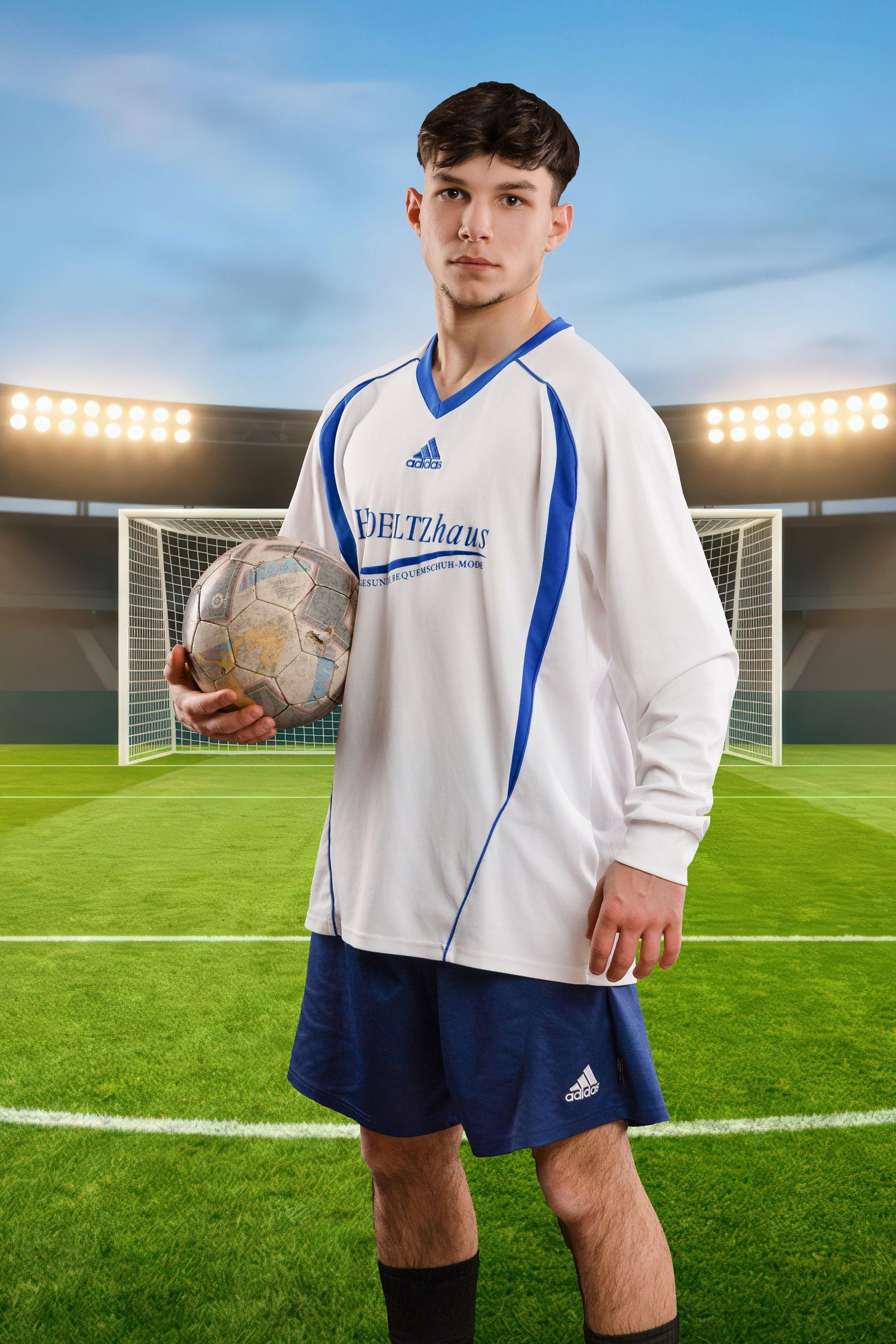 Soccer player in white and blue uniform holding a ball on field with goal post and stadium lights in background.