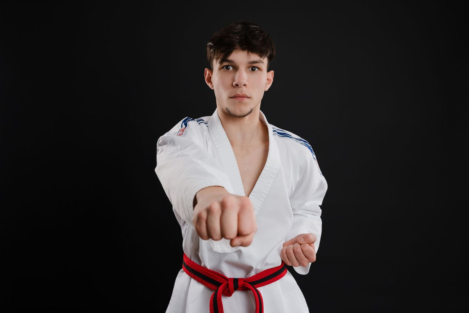 Martial artist in white karate uniform with red belt performs punch stance against dark background.