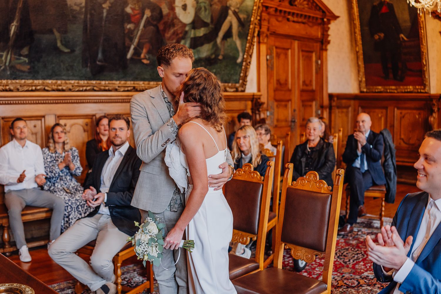 A couple shares a romantic kiss during their wedding ceremony in an ornate historic room with wooden pews and guests.