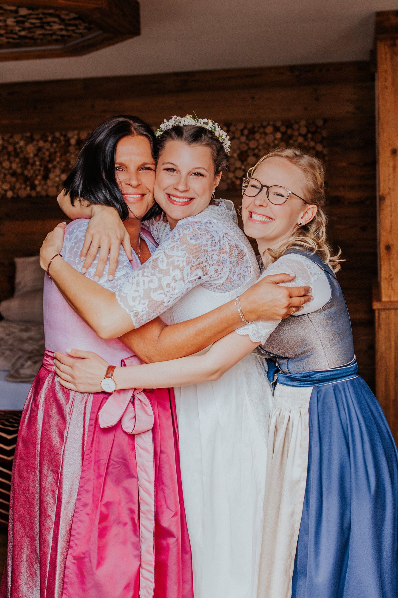 A group in colorful dirndl dresses share warm embraces in a rustic wooden setting.