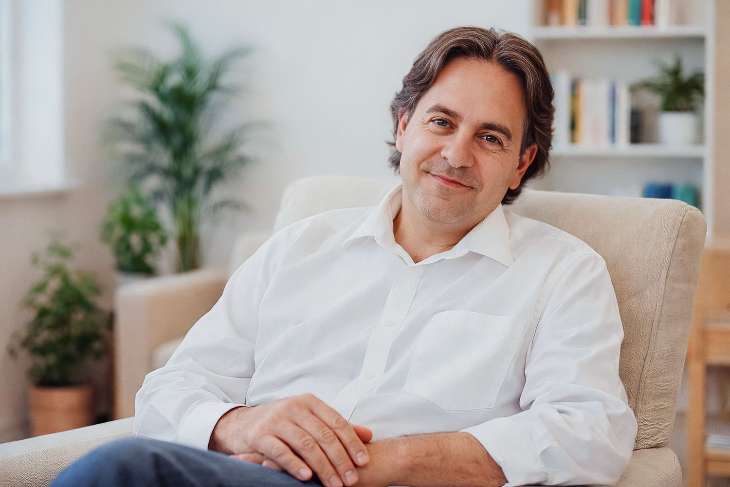 Professional wearing a white button-down shirt sits casually in a modern office space with bookshelves and plants.