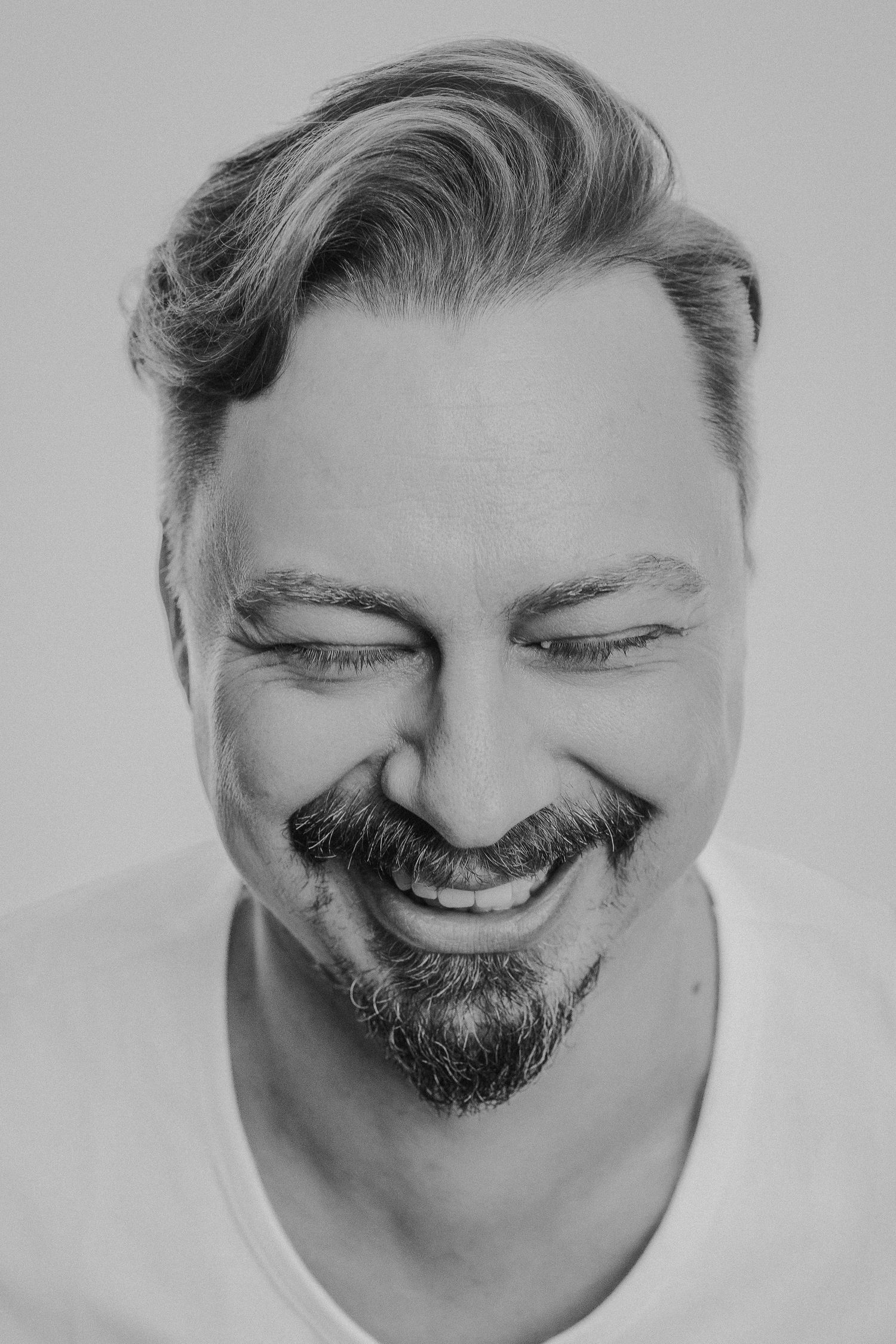 Black and white portrait photograph showing a joyful smile and neatly groomed facial hair against a plain background.