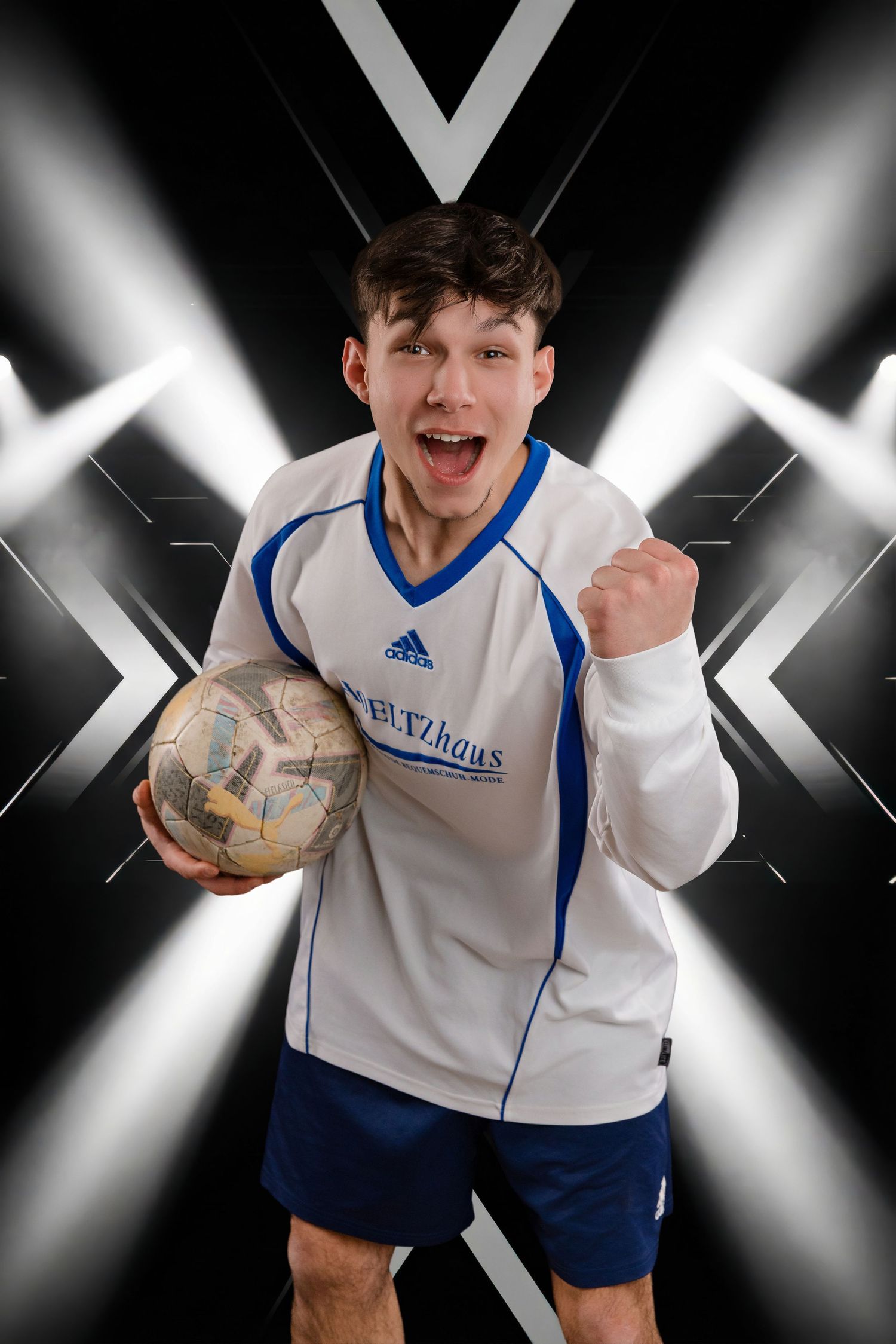 Excited athlete in white and blue uniform holds soccer ball against dramatic black background with crossing spotlights.