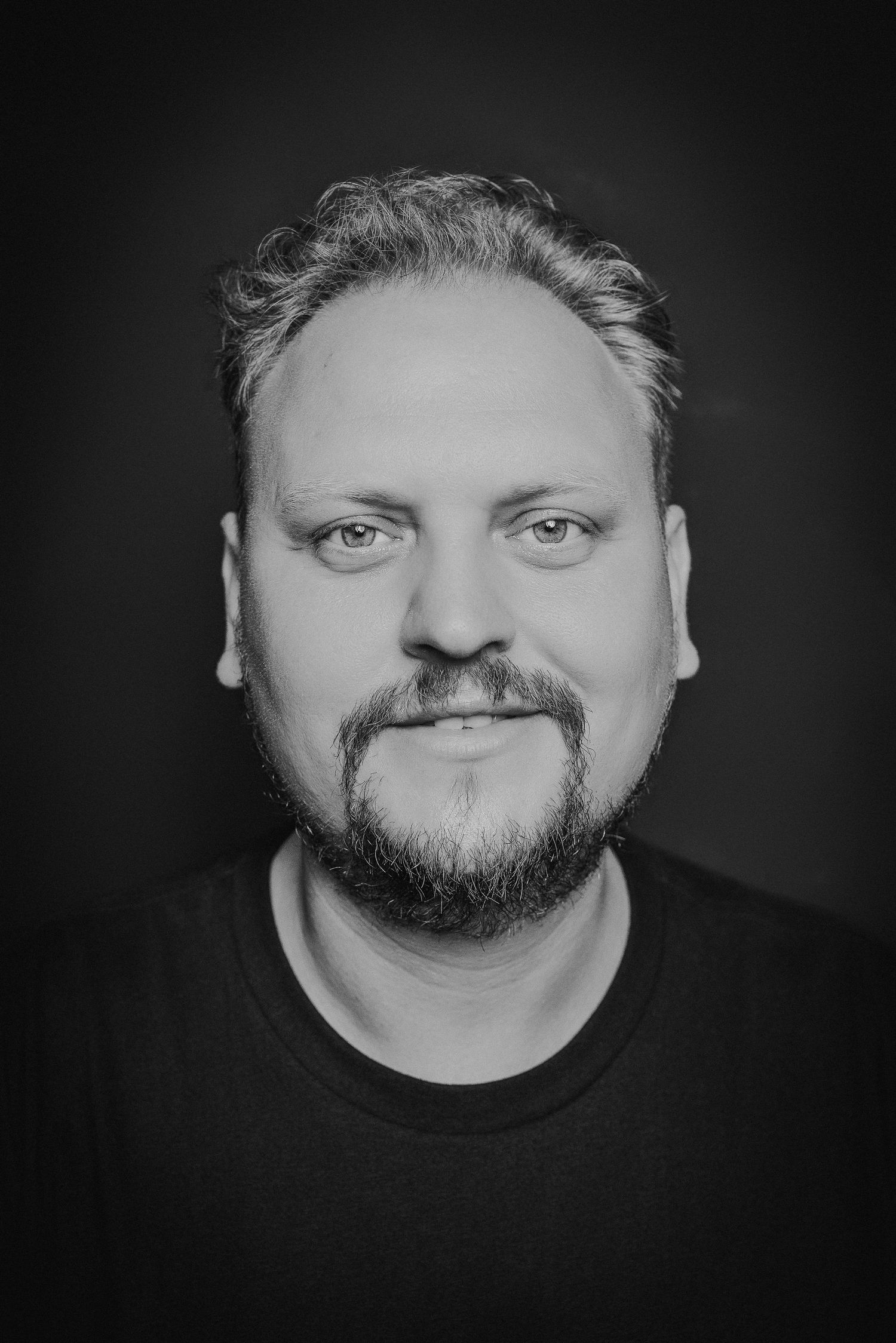 Janec selbst - der ein schwarzes T-Shirt trägt, zweigt sich lächelnd im Pure Portrait mit Blick direkt zum Betrachter.