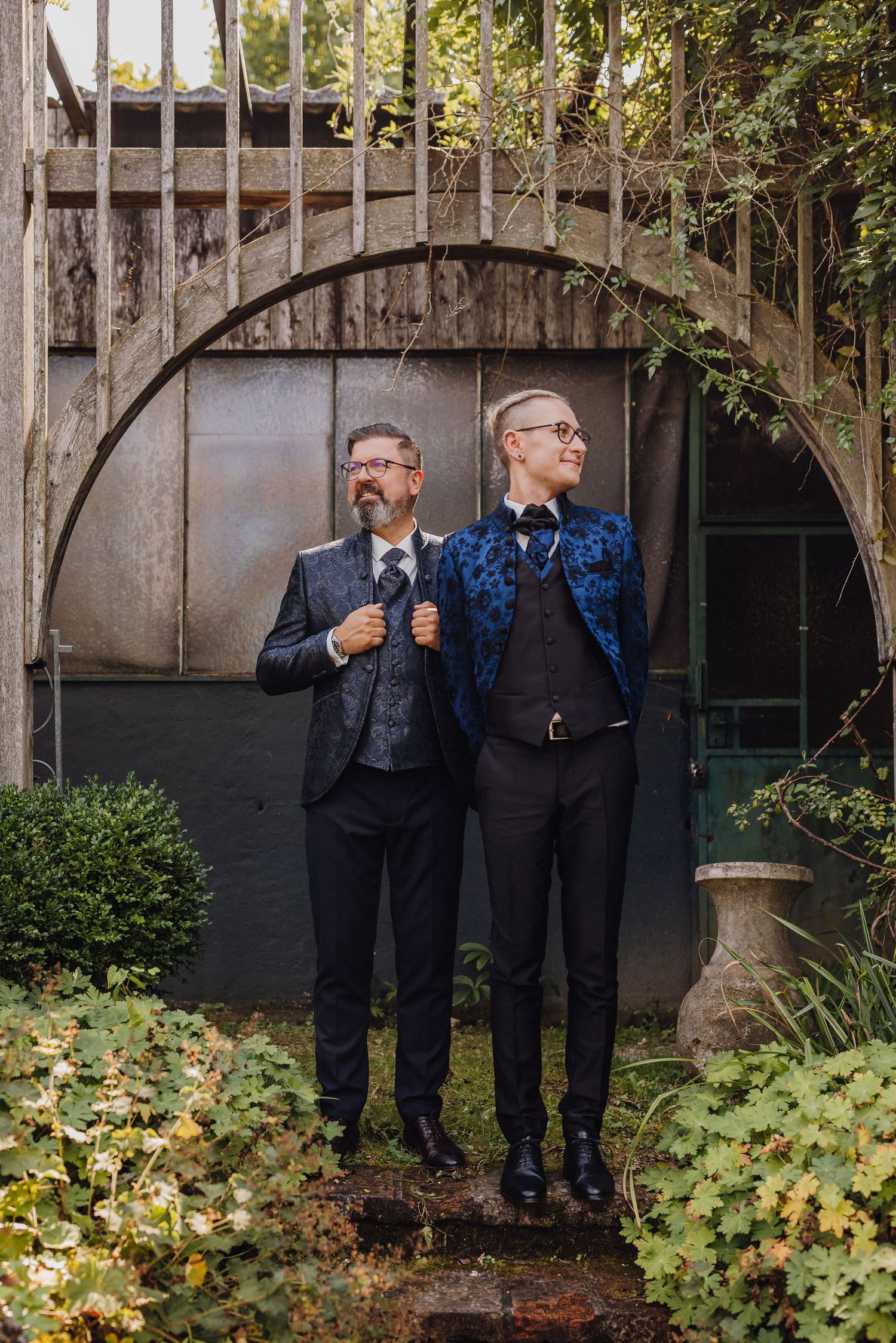 Two people in formal attire stand under a rustic garden archway wearing dark suits with one in a blue patterned jacket.