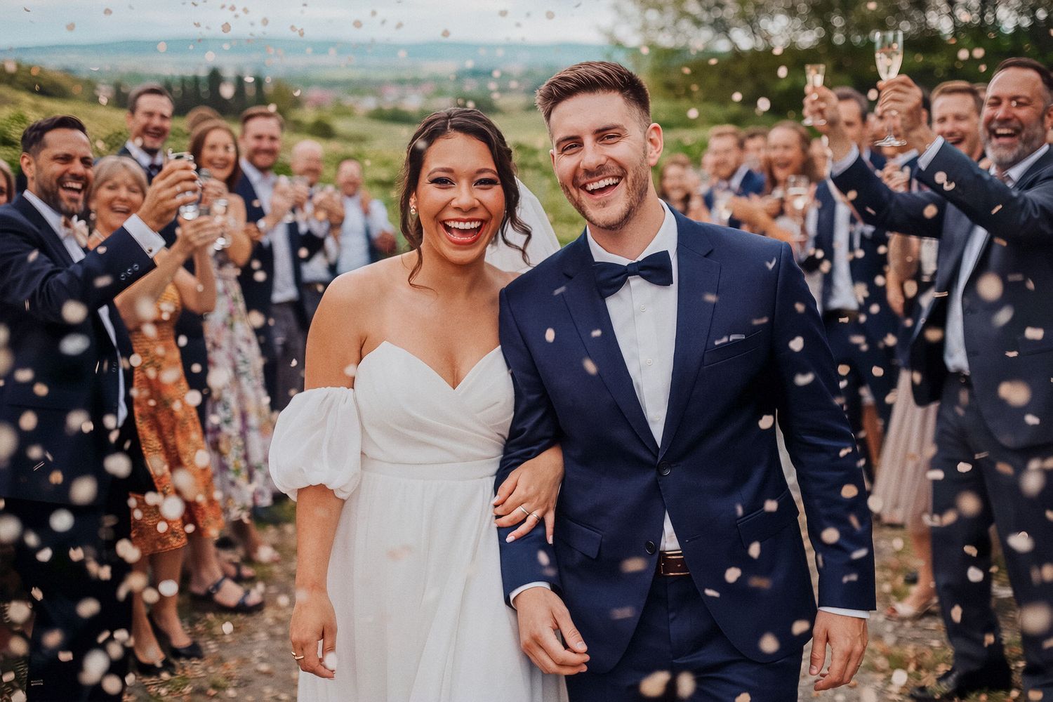 A joyful newlywed couple walks through confetti while wedding guests celebrate in the background.