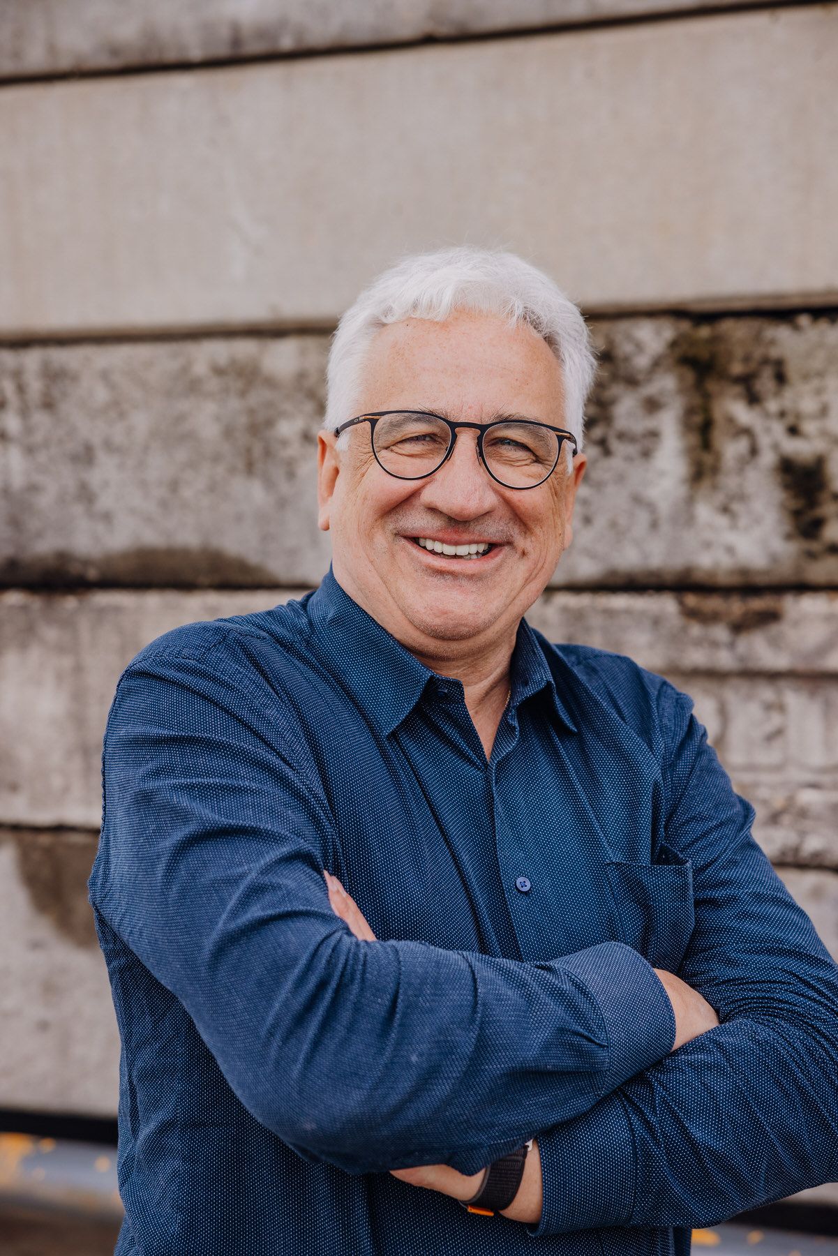 A person in glasses and a navy blue button-down shirt stands with crossed arms against a textured wall.