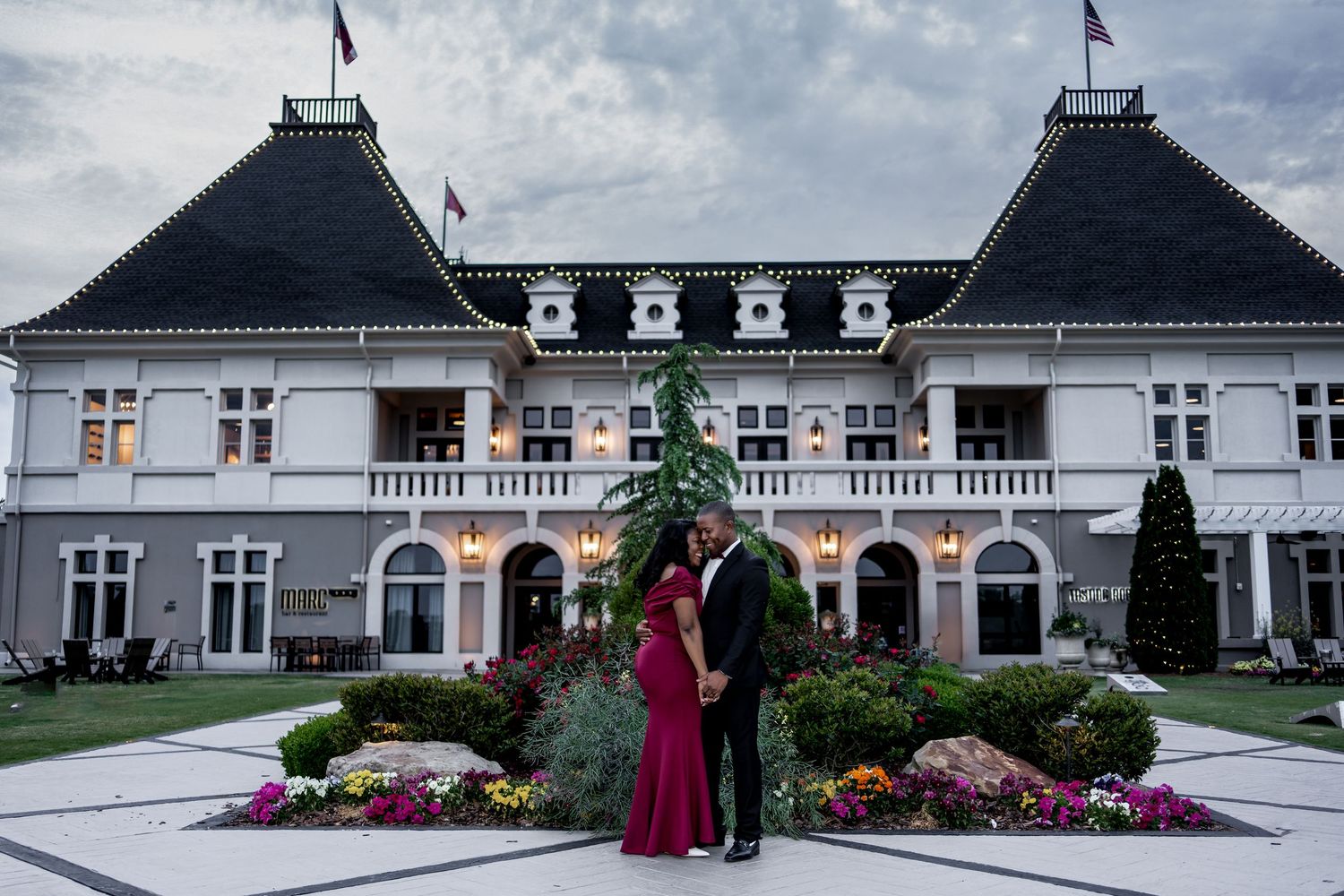 Elegant couple stands in front of a grand white mansion with twin spires and colorful flower gardens at dusk.