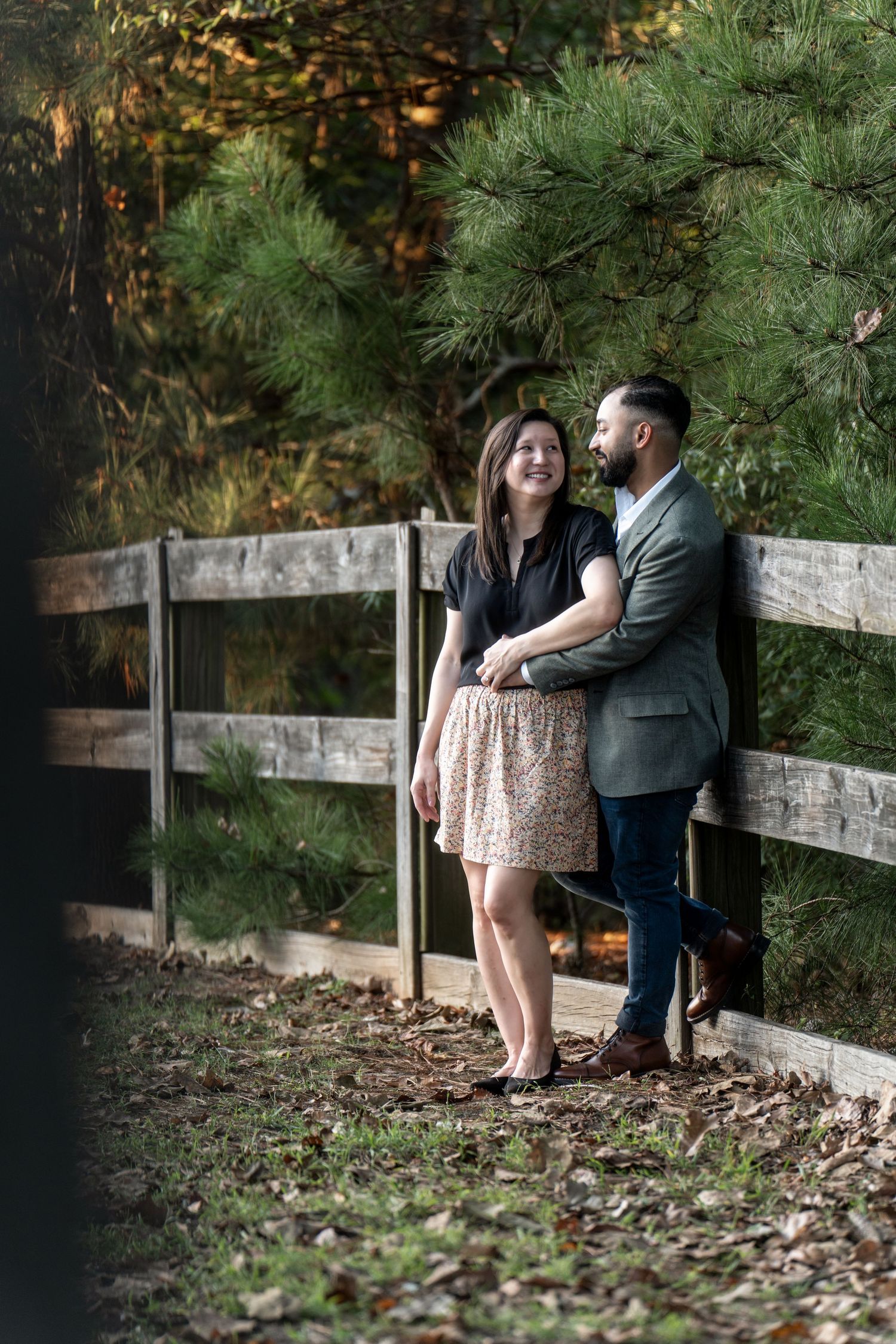 A couple shares a romantic moment along a rustic wooden fence surrounded by pine trees in a natural outdoor setting.