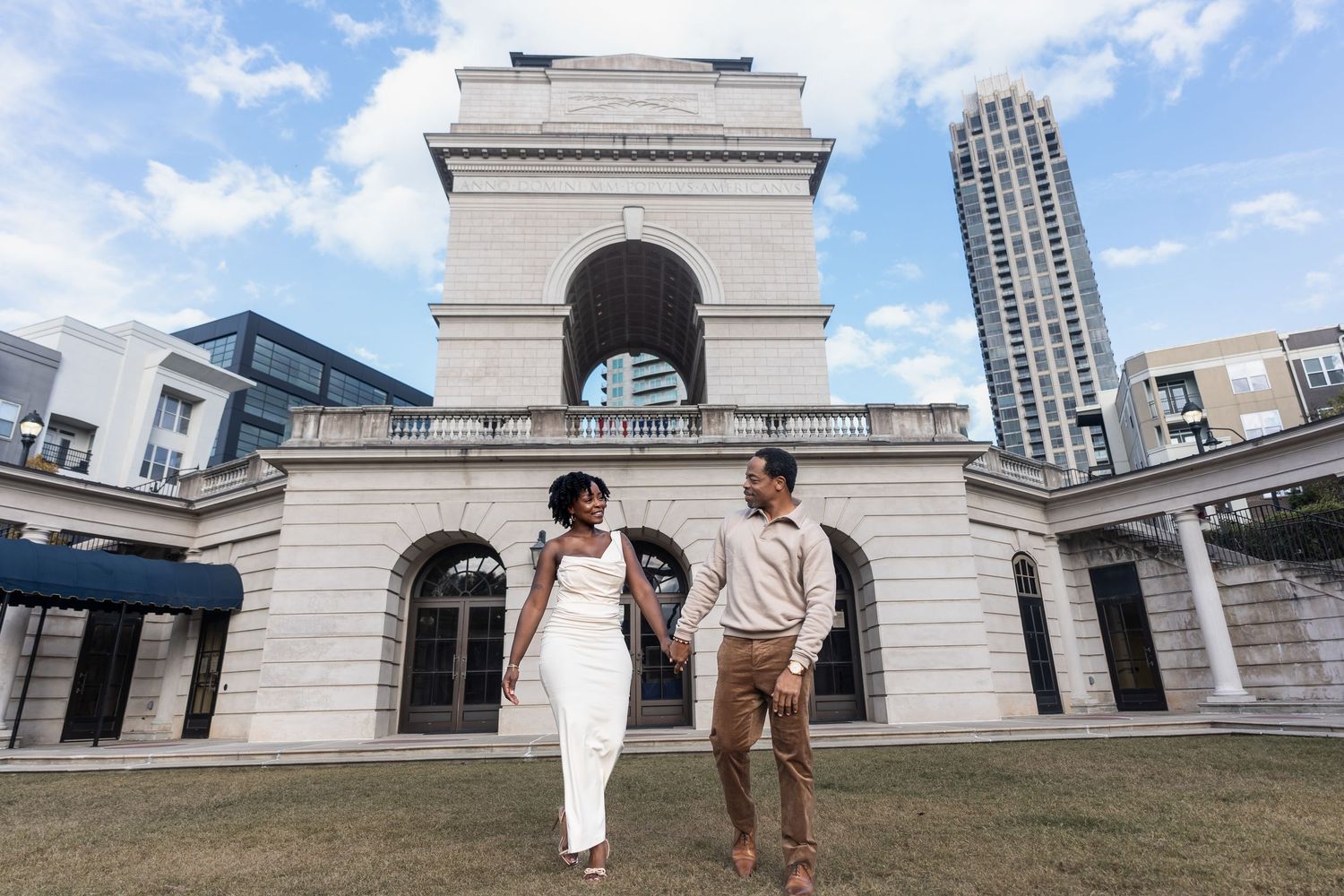A couple shares a romantic moment in front of a white stone building with arched columns on a sunny day.