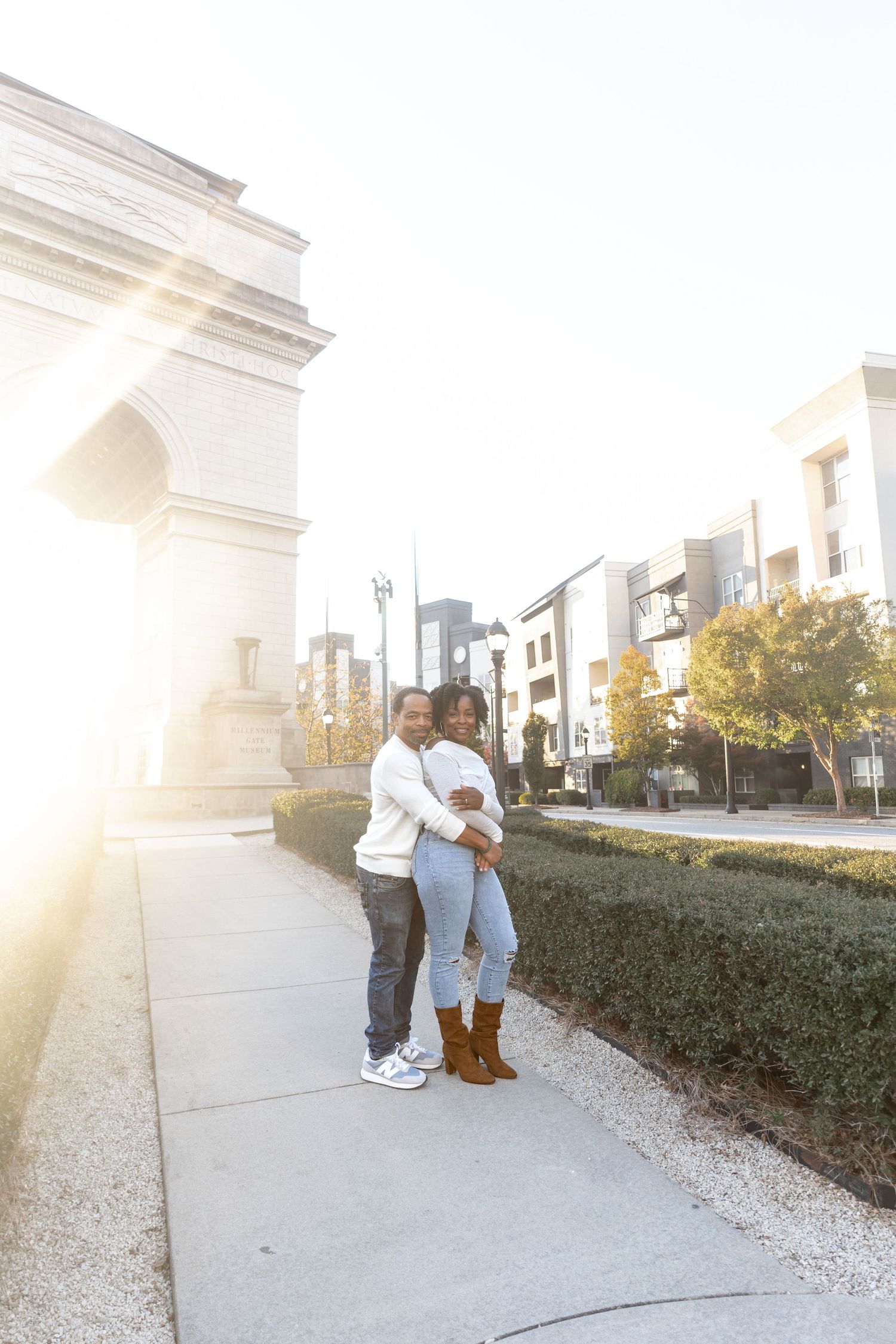 A couple embraces during a romantic sunset photoshoot in front of a white building with landscaping.