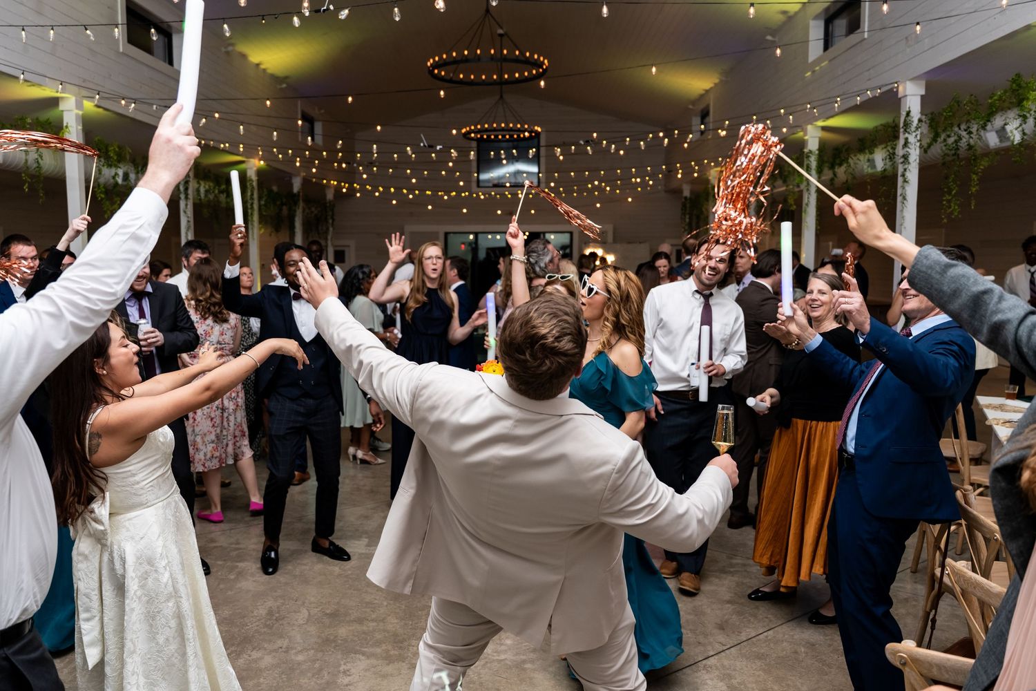 Wedding reception guests celebrate and dance with arms raised under twinkling lights at an indoor venue.