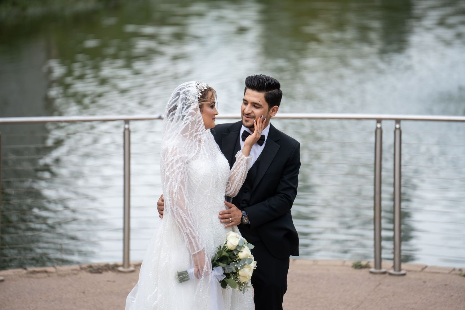 Newlywed couple shares romantic moments by a lakeside railing during their wedding photoshoot.