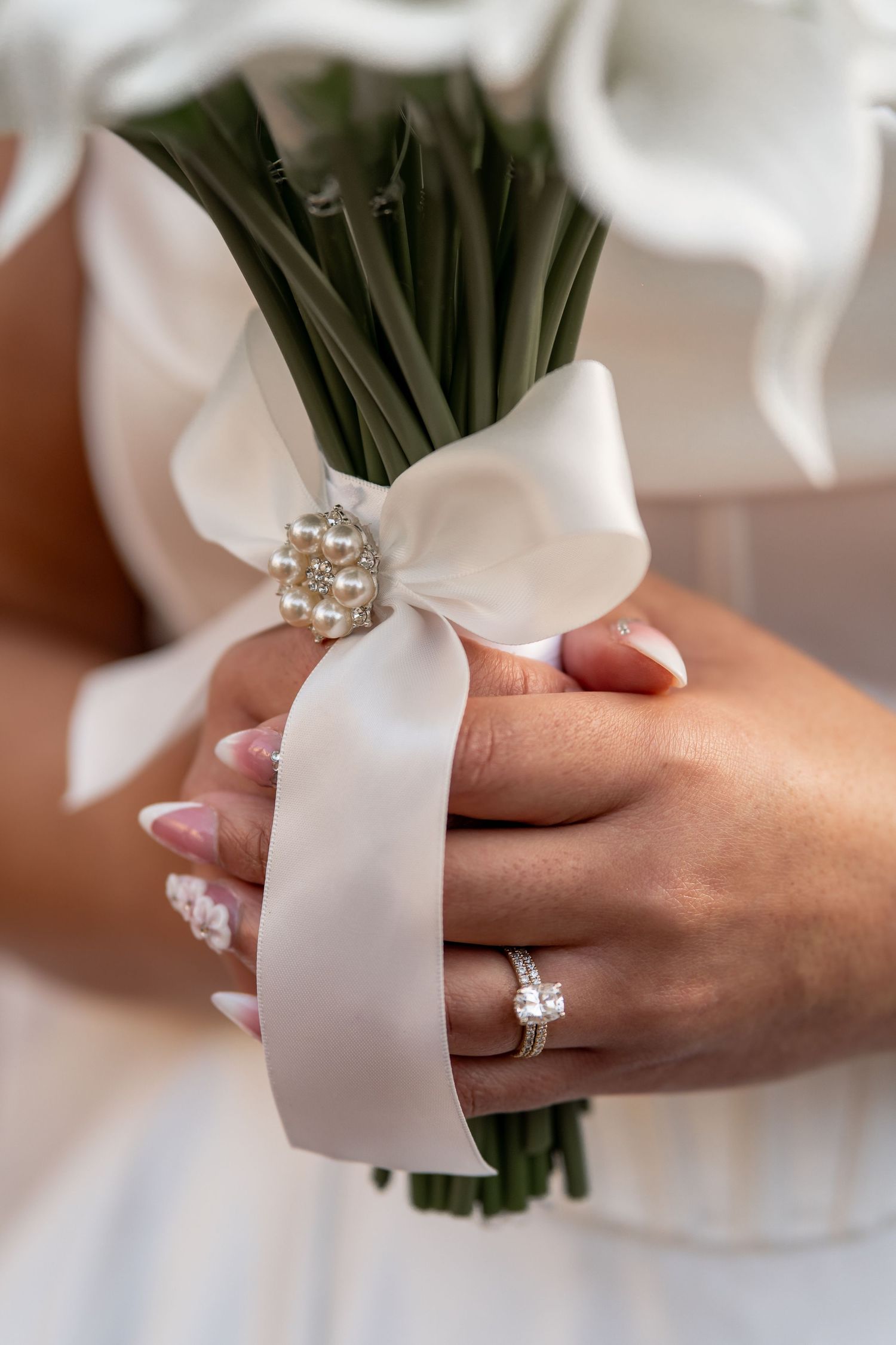 Close-up of hands holding a white ribbon-wrapped wedding bouquet with a diamond engagement ring visible.